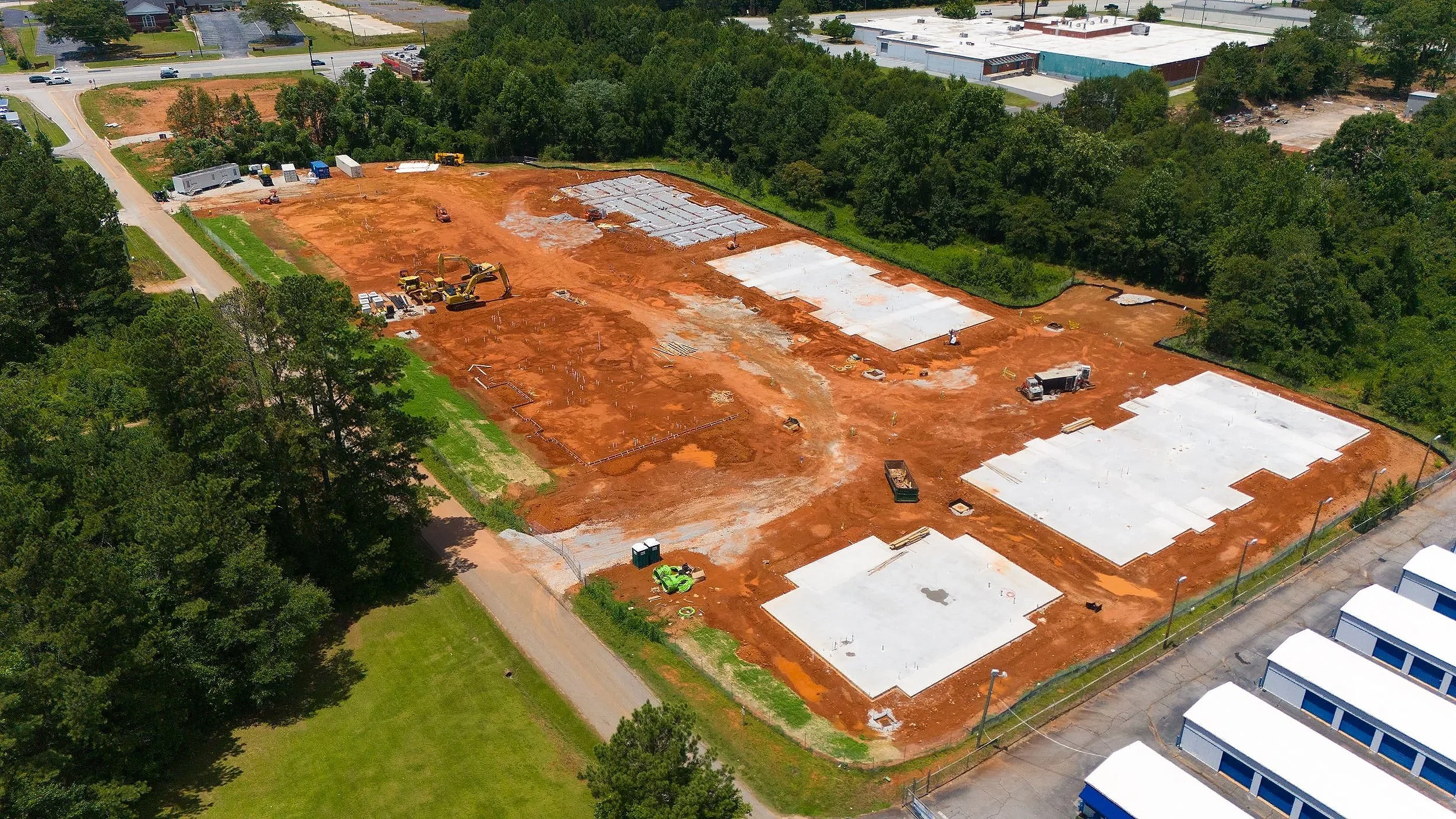 Construction site with several building foundations and machinery, dirt ground, surrounded by trees and a partially paved road.