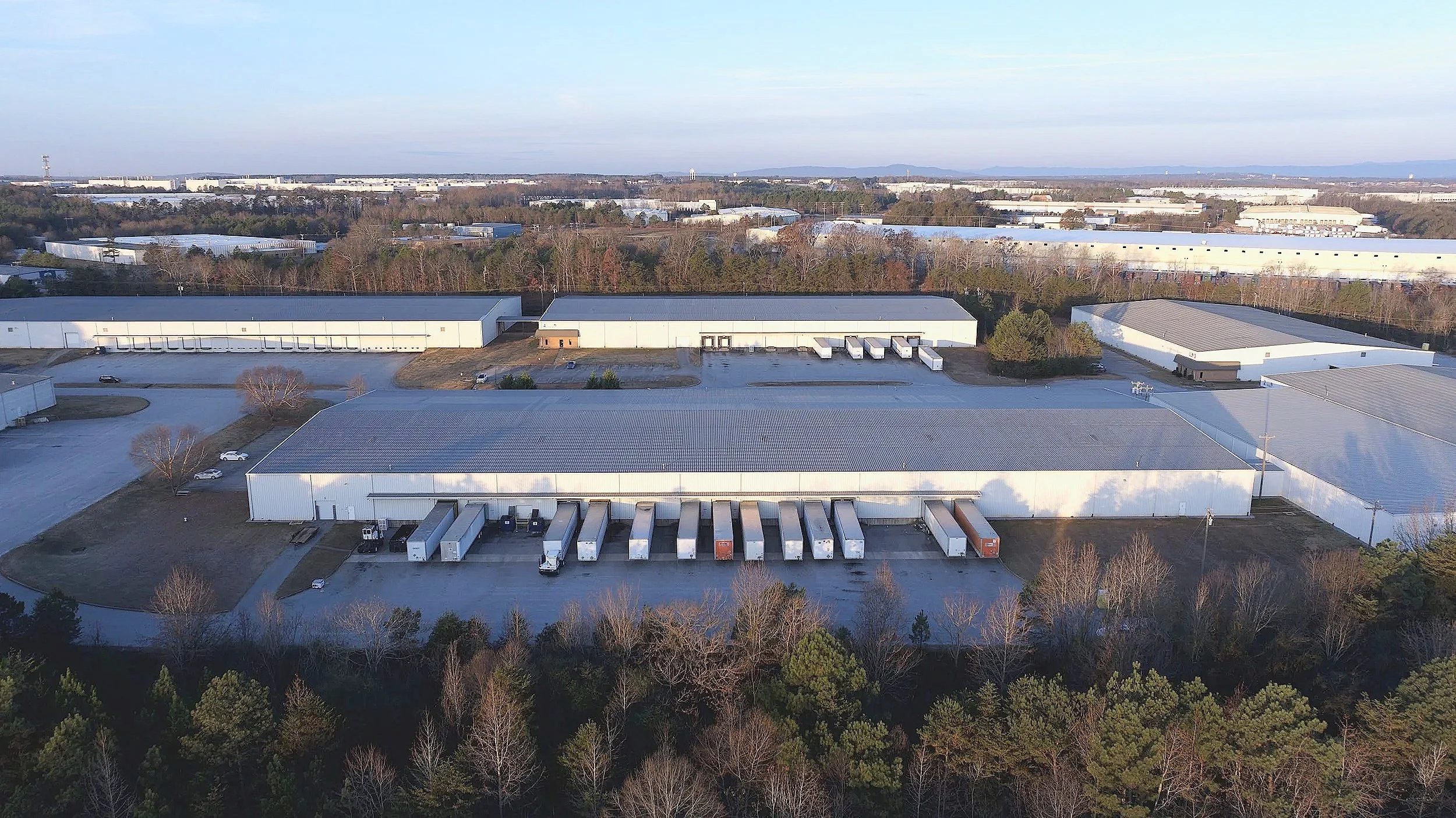 Aerial view of an industrial warehouse complex with multiple large white buildings, loading docks with trucks, surrounded by trees and parking areas, under a clear sky.