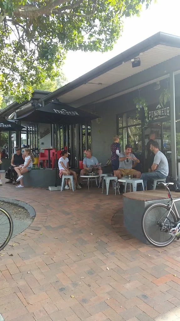 People sitting and standing outside a cafe or restaurant with outdoor seating, bicycles parked nearby, greenery overhead, and a building with large windows.