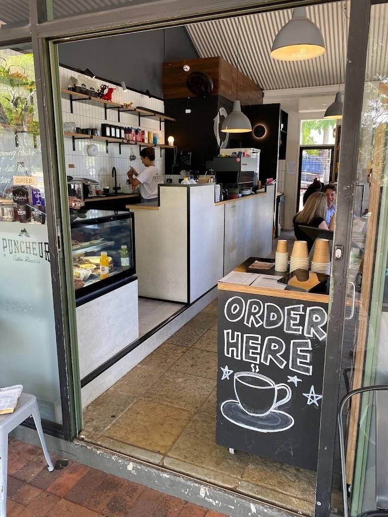 Inside a coffee shop with a counter, a barista preparing drinks, and customers seated at tables. There is a chalkboard sign outside that says 'Order Here' with a drawing of a steaming cup of coffee.