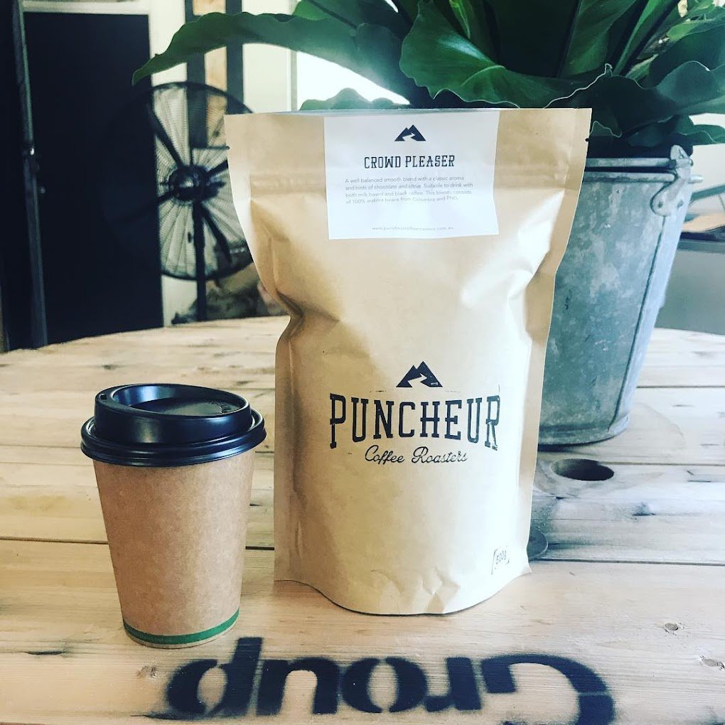 A paper coffee cup with a black lid placed on a wooden table, next to a bag of coffee labeled 'Punchieur Coffee Roasters' and a large potted plant in the background.