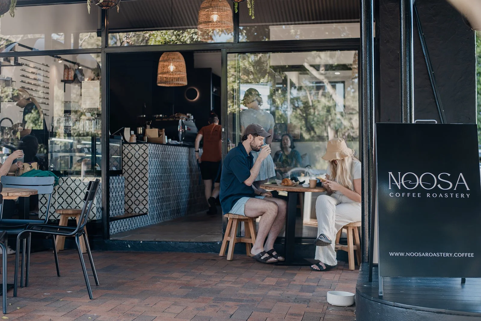 A man and woman sit at a small table outside a coffee shop, drinking coffee and using their phones. The coffee shop sign reads 'NOOSA COFFEE ROASTERY'. The interior of the shop is visible through the large window, with baristas working behind the cou