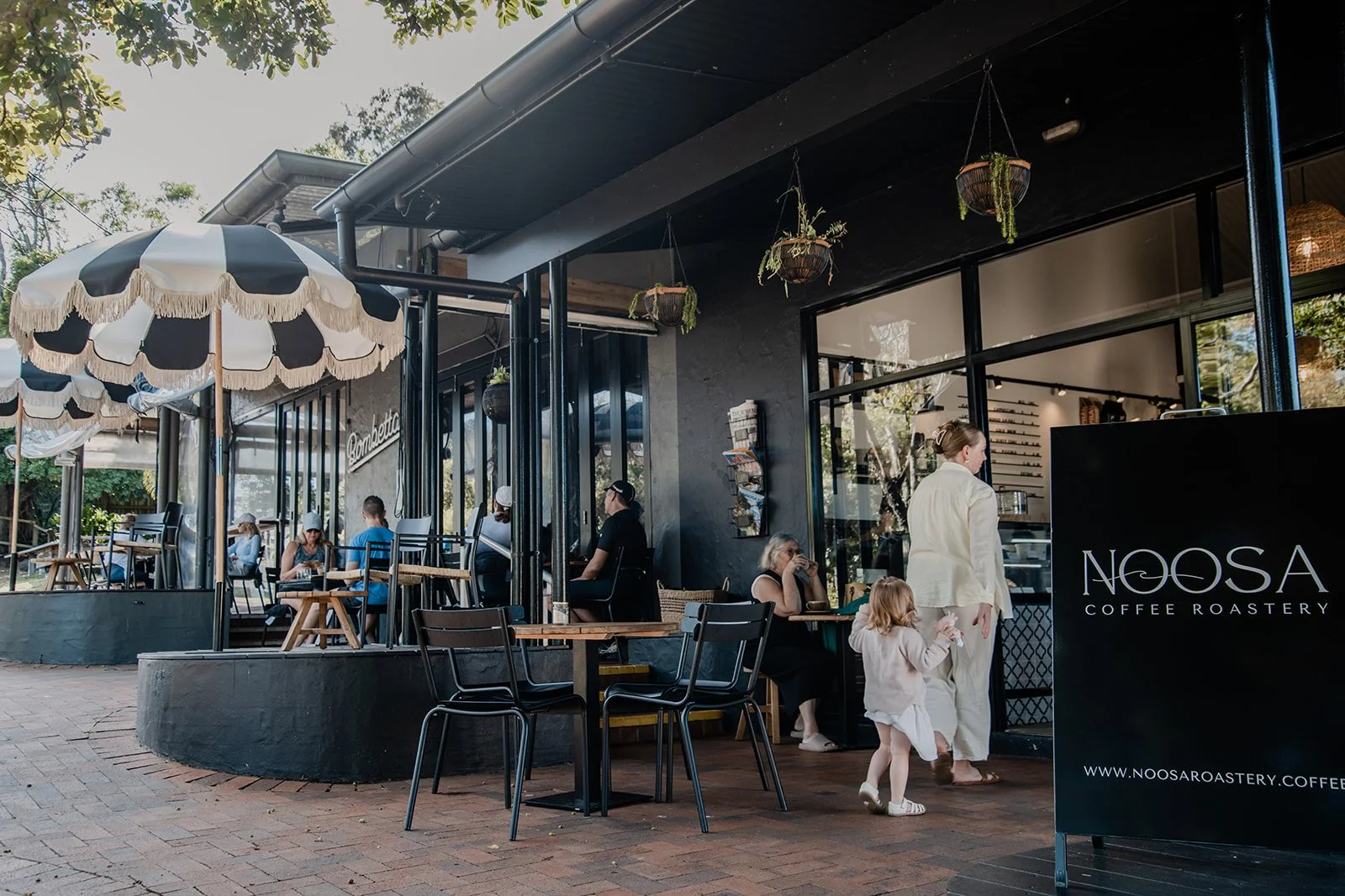 Outside view of Noosa Coffee Roastery with seating area, umbrellas, and customers, including a woman and child entering the cafe.