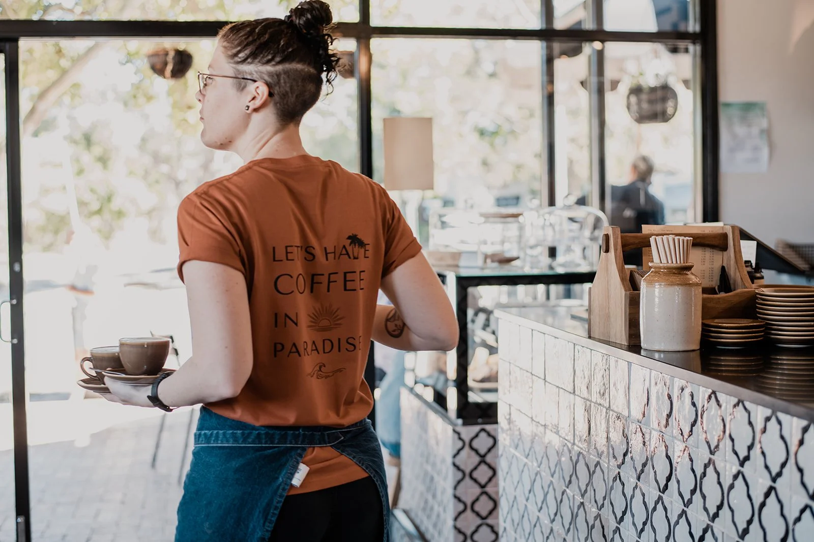 A barista holding two coffee cups stands behind the counter of a cafe, with a laptop and coffee cups on the counter, and a person outside visible through large windows in the background.