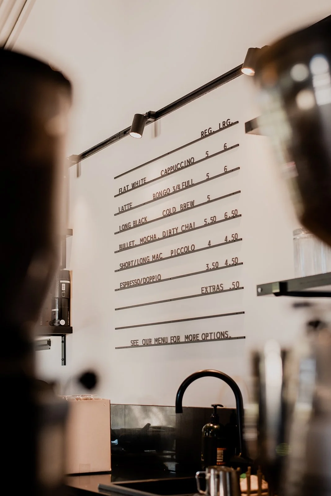 Coffee shop menu board with various drink options and prices, partially visible behind the counter with coffee brewing equipment.