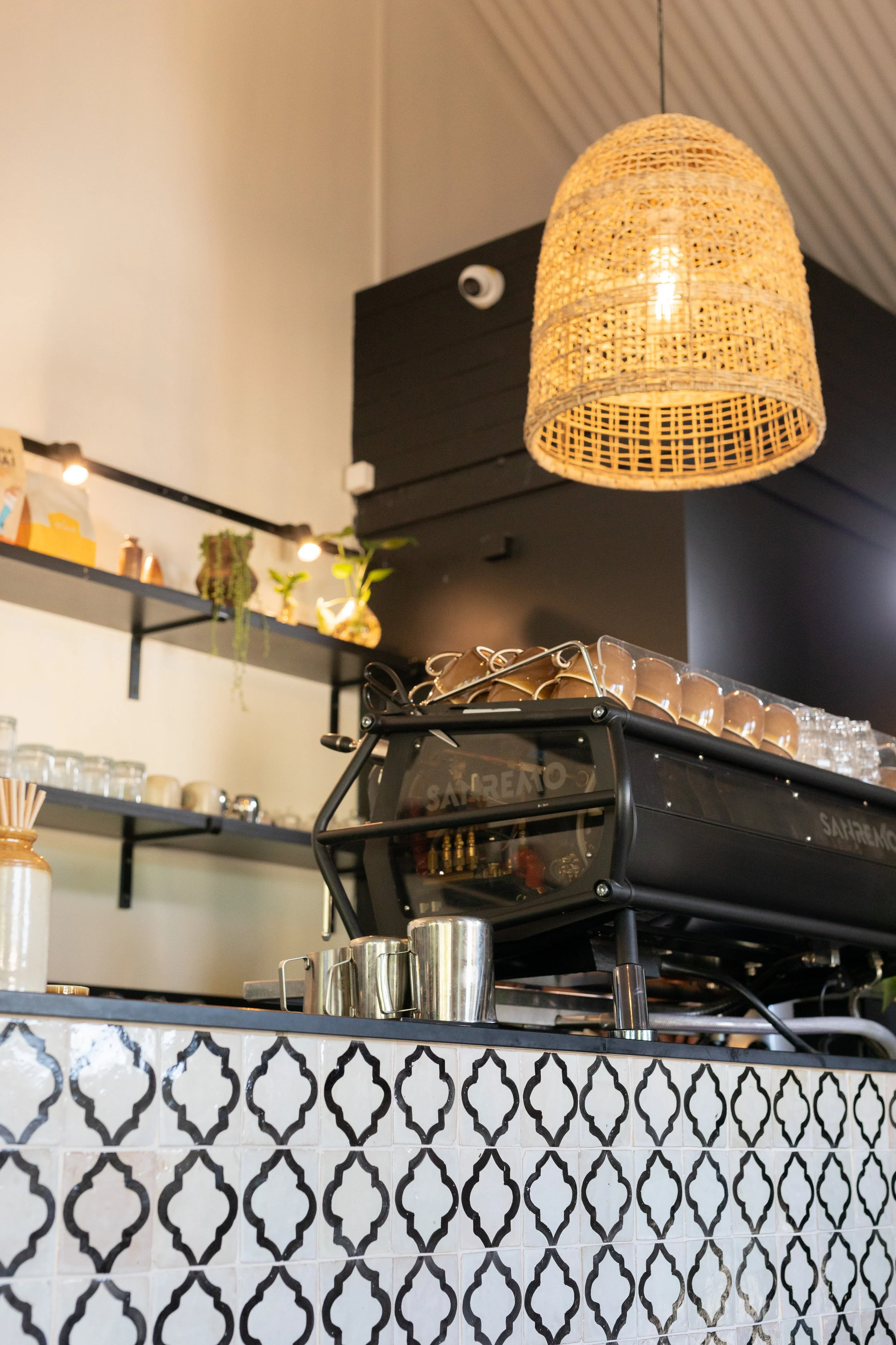 Coffee shop counter with a black espresso machine on a tiled counter, overhead rattan pendant light, shelves with glassware and plants in the background.