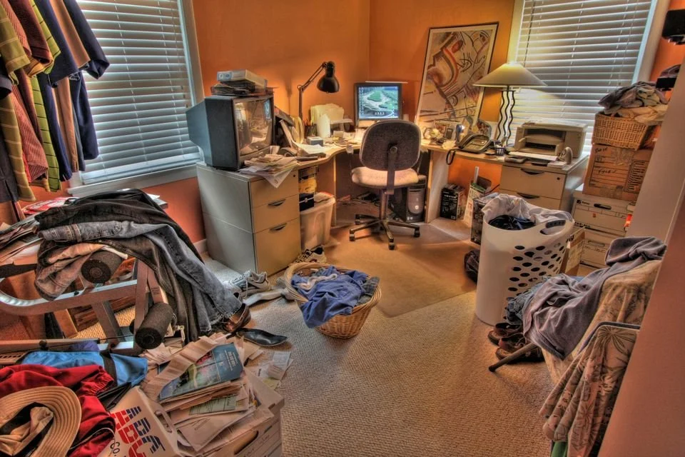 Cluttered home office with laundry, papers, and miscellaneous items around a desk with a computer, lamp, and chair.