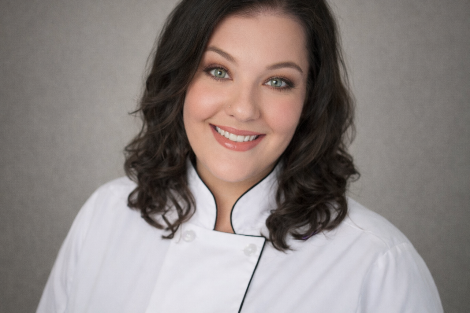 Portrait of a woman with long, curly dark hair and green eyes, smiling, wearing a white chef's coat with black trim, against a gray background.