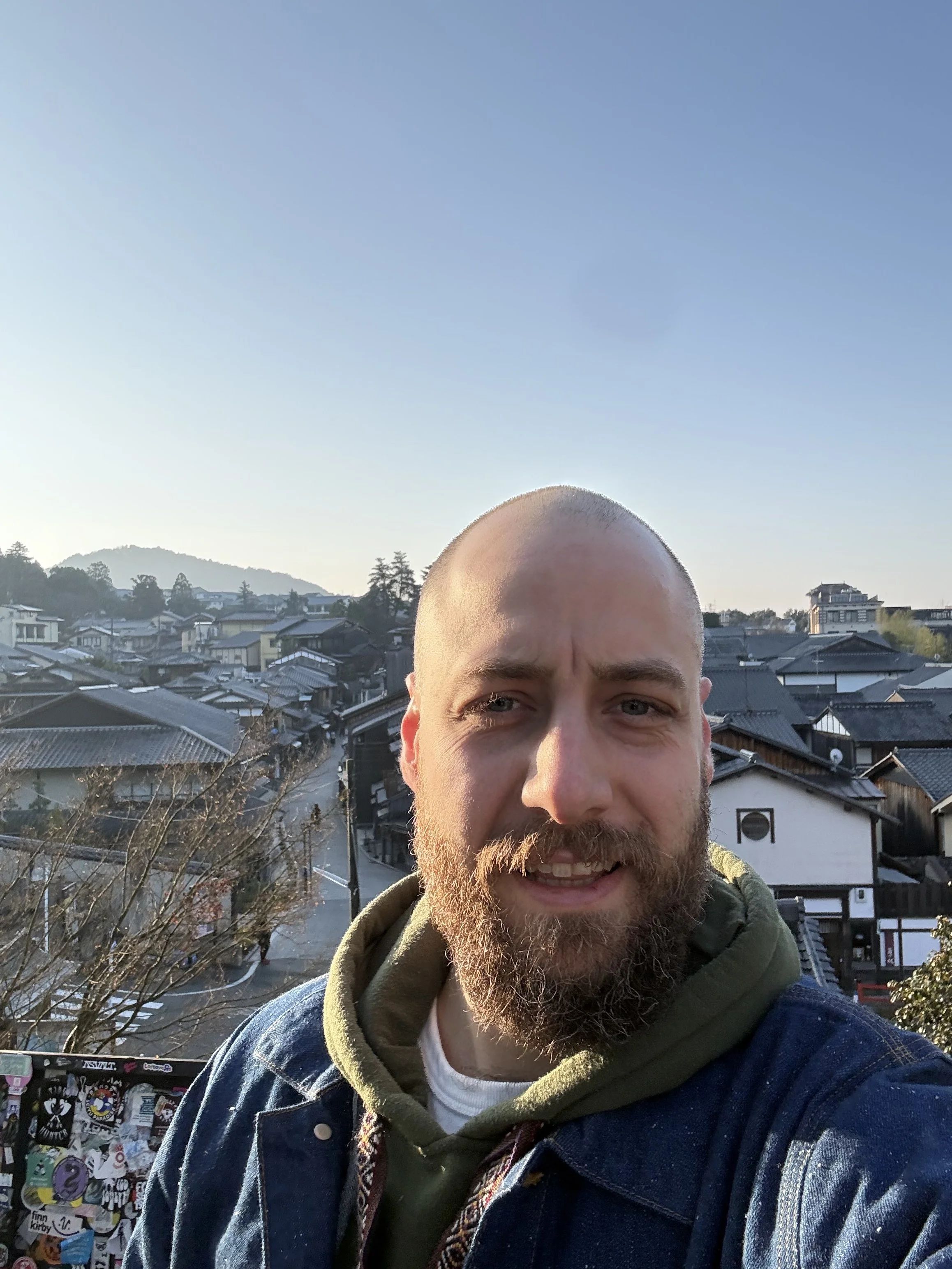 A man with a beard and bald head taking a selfie outdoors in front of a traditional Japanese town with tiled roofs and a mountain in the background.