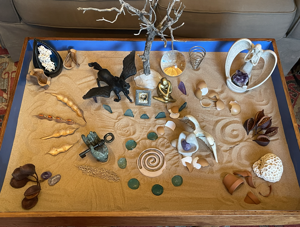 A desert-themed decorative arrangement on a wooden table with sand, shells, stones, crystals, a tree sculpture, a small statue, and various artistic objects.