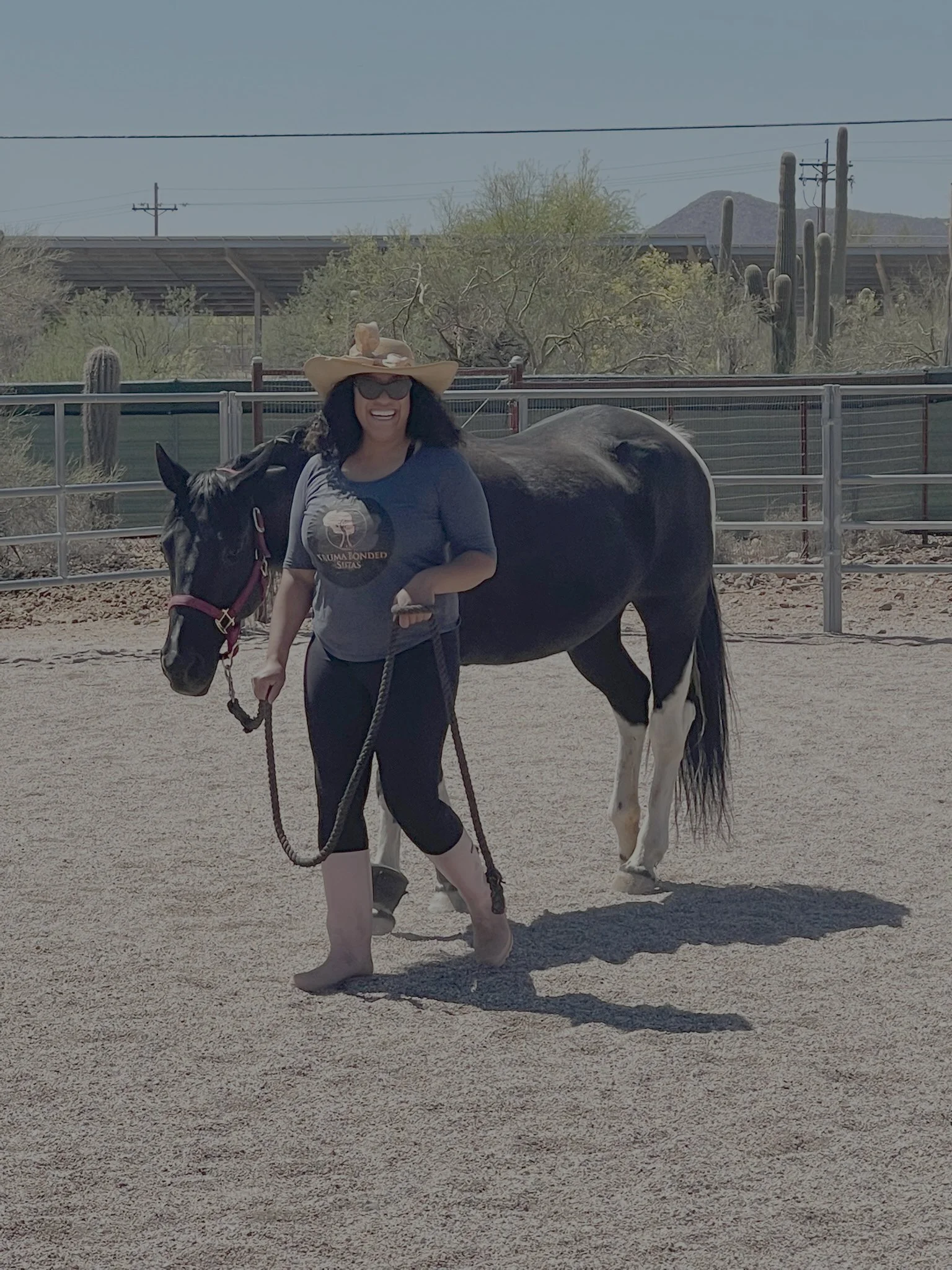 A woman wearing a large straw hat, sunglasses, a gray T-shirt, and black leggings is walking a black-and-white horse in an outdoor setting with desert trees, cacti, and mountains in the background.