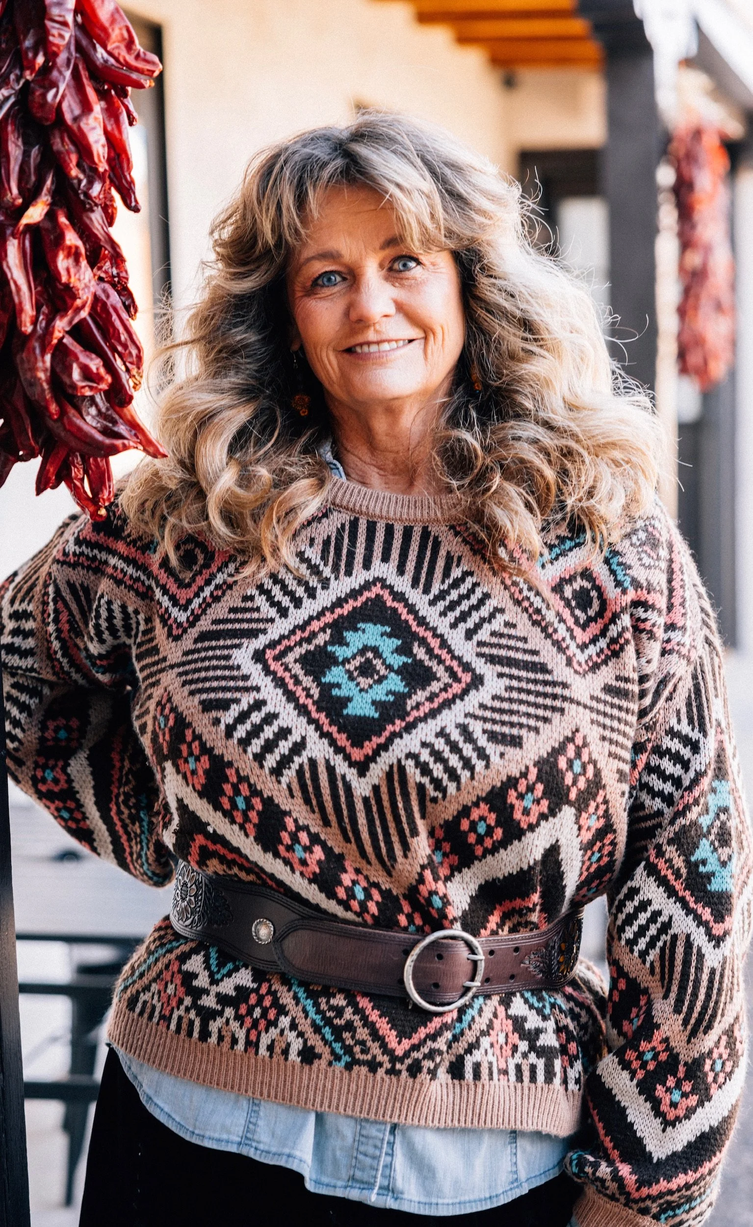 A woman with long wavy blonde hair and blue eyes standing outdoors near a wooden structure with hanging chili peppers. She is wearing a colorful patterned sweater with a belt and a light shirt underneath, smiling at the camera.