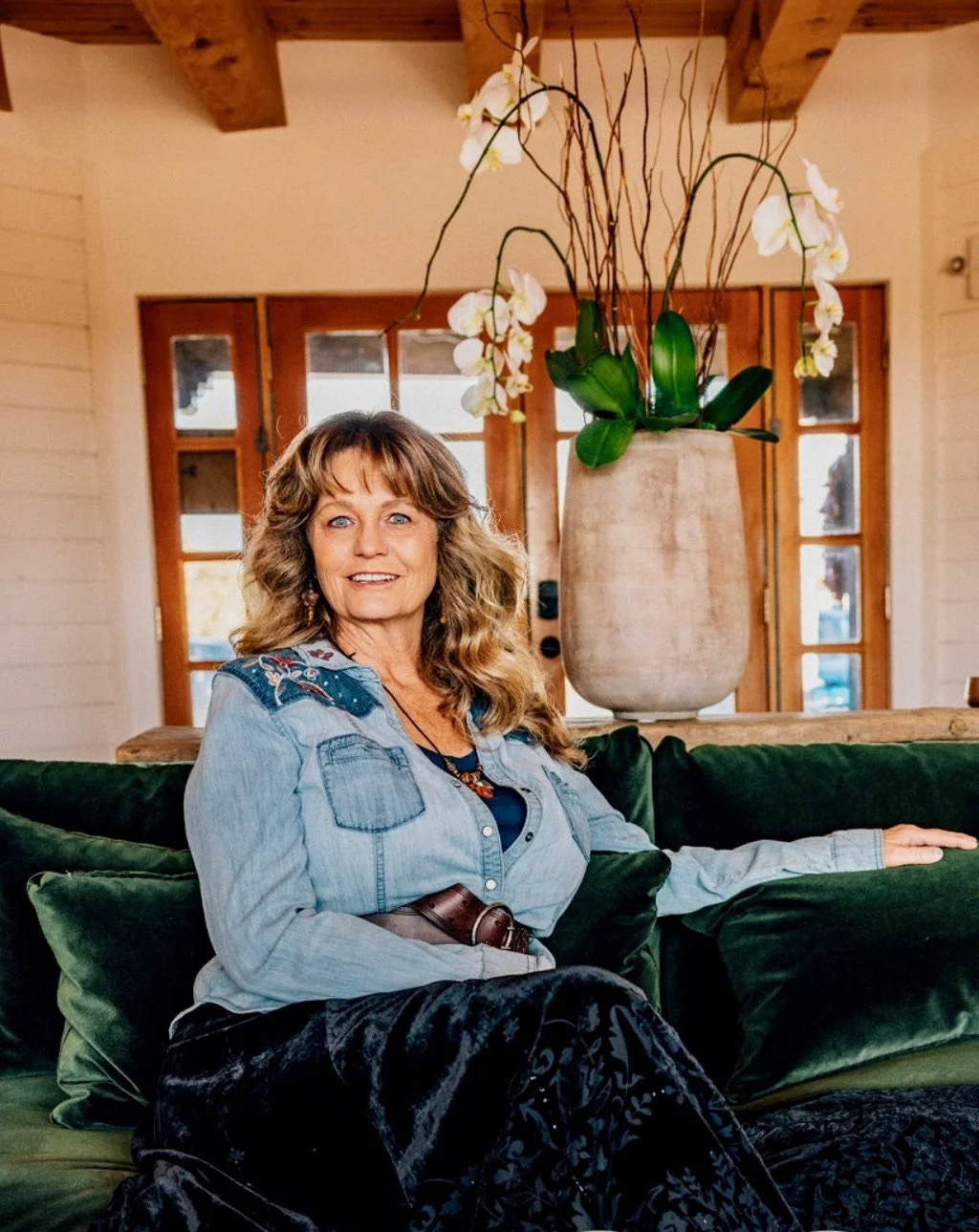 A woman with wavy blonde hair wearing a denim shirt with embroidered floral details, sitting on a green velvet couch in a cozy room with large glass doors, a wooden ceiling, and a large vase with white orchids and green leaves behind her.