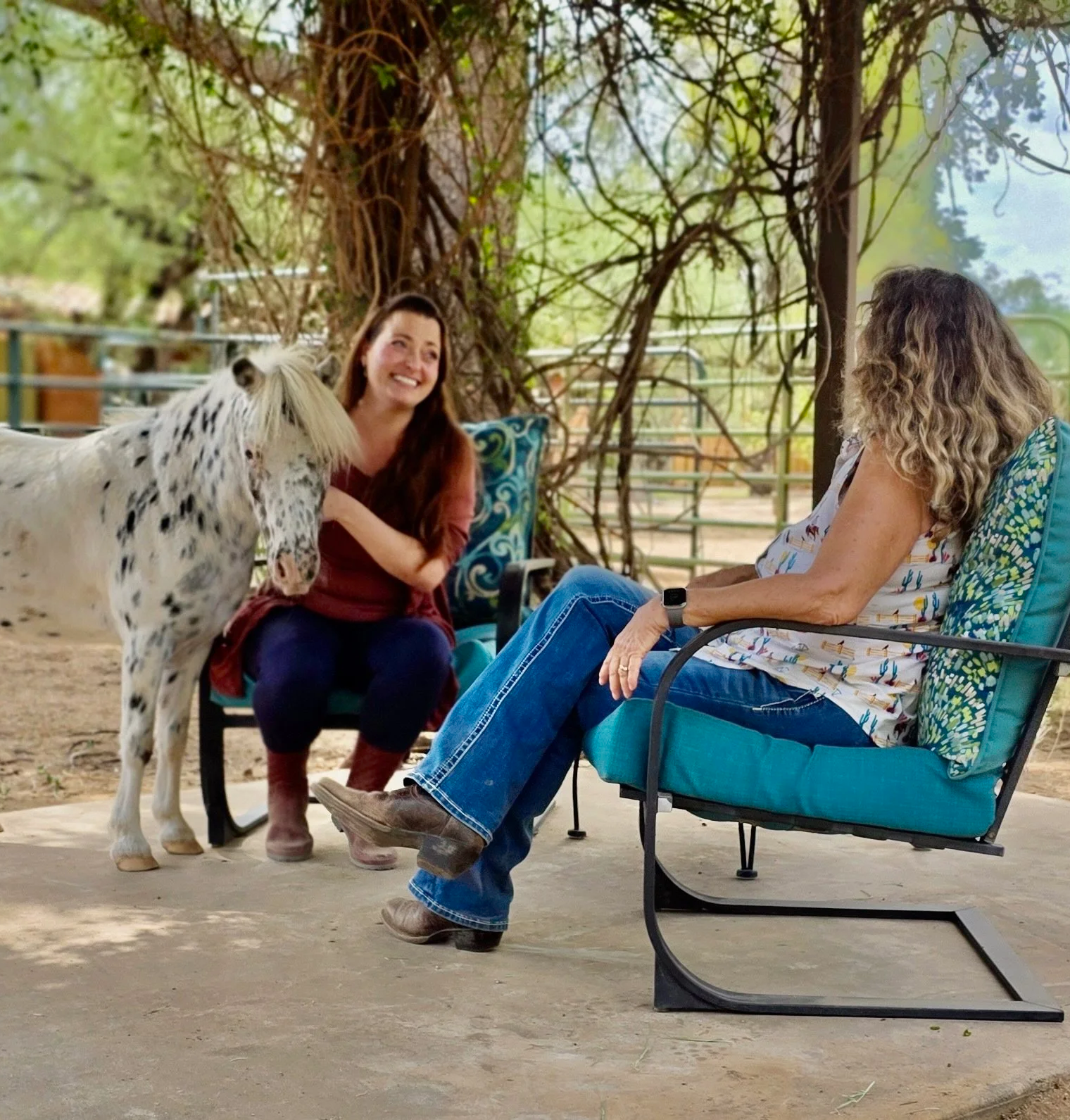 Two women sitting outdoors under a large tree, with a small white horse with black spots on its face. One woman has long brown hair and is smiling, while the other has curly blonde hair and is sitting in a teal chair, wearing a patterned blouse.