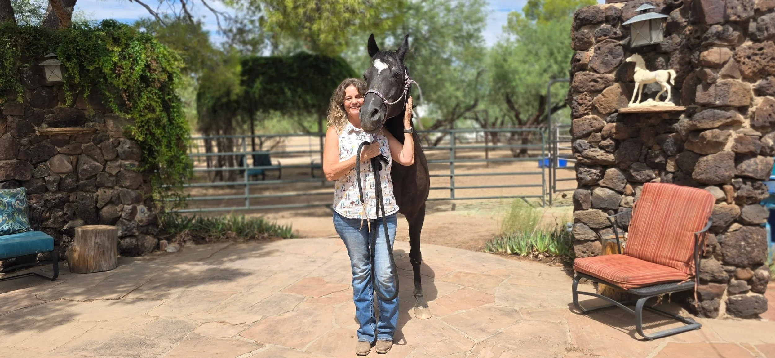 A woman smiling and holding a black horse's bridle outdoors on a stone patio. Behind her is a wrought iron fence, with trees and a clear sky in the background. The patio is decorated with outdoor furniture, including a blue cushioned chair, a striped cushioned chair, and a small tree stump.