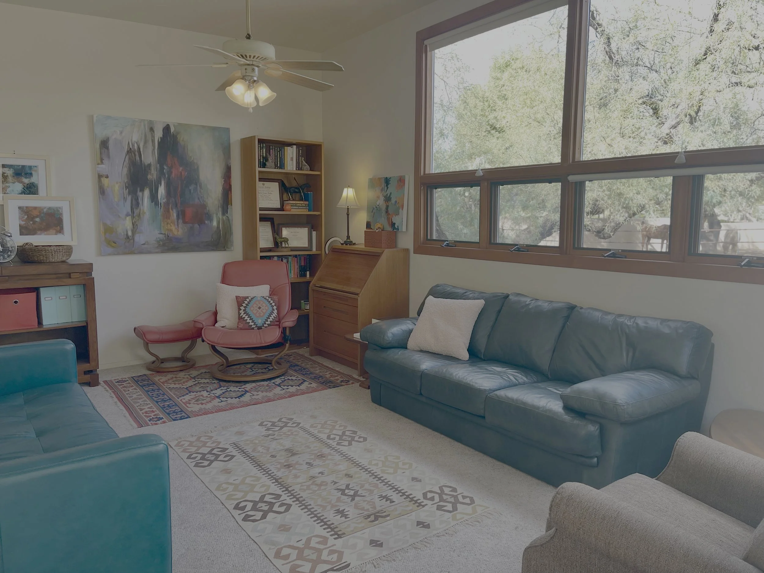 Living room with large windows, green leather sofa, beige armchair, red armchair with pillow, bookshelf, colorful paintings, ceiling fan, and a rug.