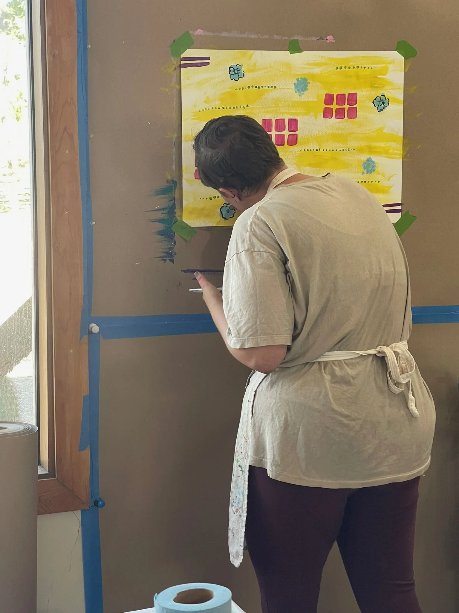 A person with short dark hair wearing a beige apron, painting on a colorful wall hang with a paintbrush in their right hand, in a room near a window with natural light.