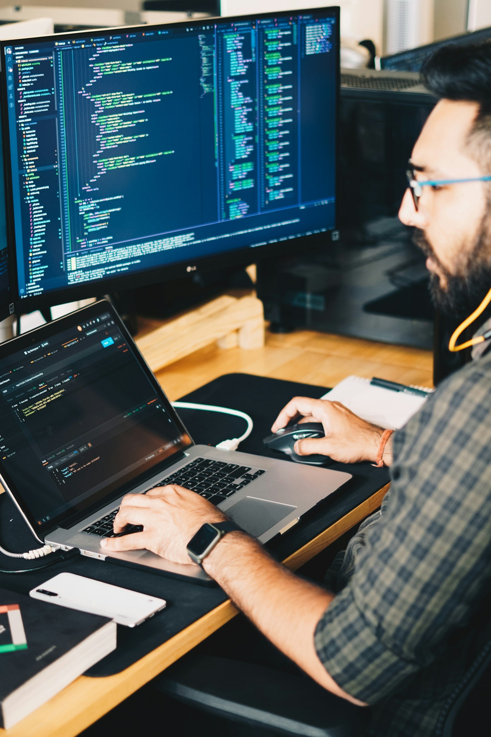 Person working on coding projects with multiple monitors and a laptop, surrounded by tech accessories on a wooden desk.