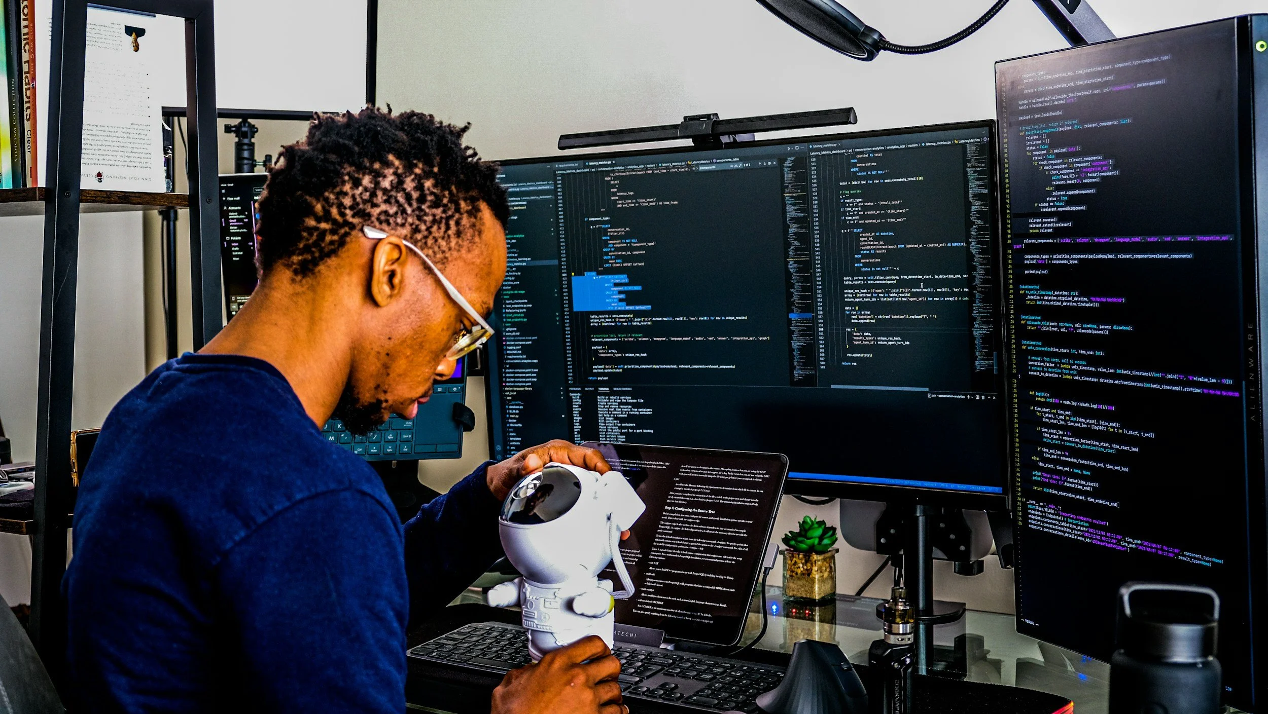 A person with glasses is working at a desk with multiple monitors displaying coding on the screens. They are holding a robot figurine.