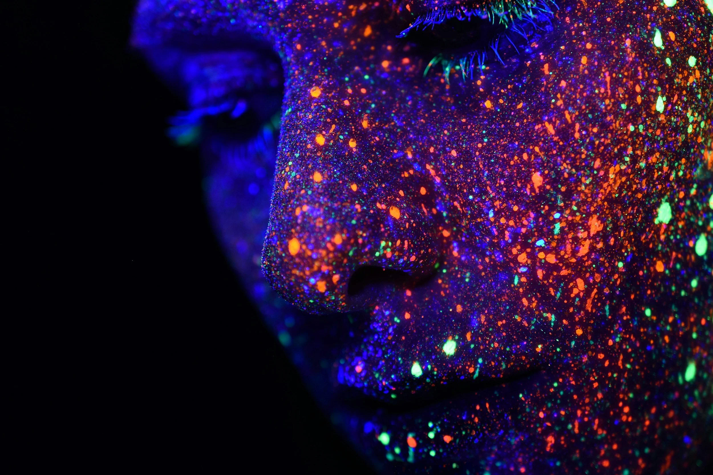 Close-up of a person's face with black light reactive neon paint, showcasing their closed eye, nose, and lips covered in colorful neon dots and splatters.