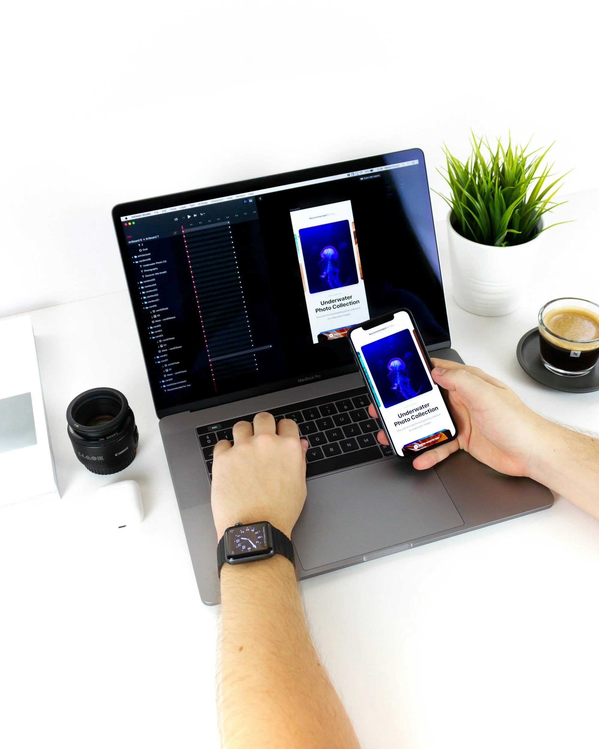 Person working on a MacBook Pro and iPhone showing a photo collection app about underwater jellyfish, with a camera lens, coffee, plant, and smartwatch on a white desk.