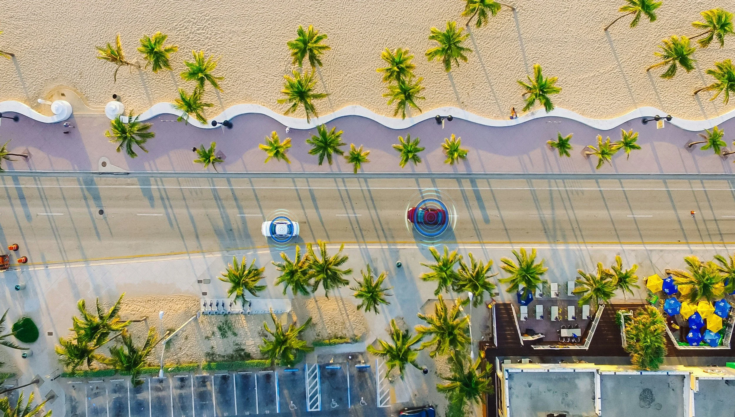 An aerial view of a beach promenade with palm trees, a sandy beach, and two people on the sidewalk. Surfboard-shaped structures line the beach, and a beachside café with yellow umbrellas is visible at the bottom right.