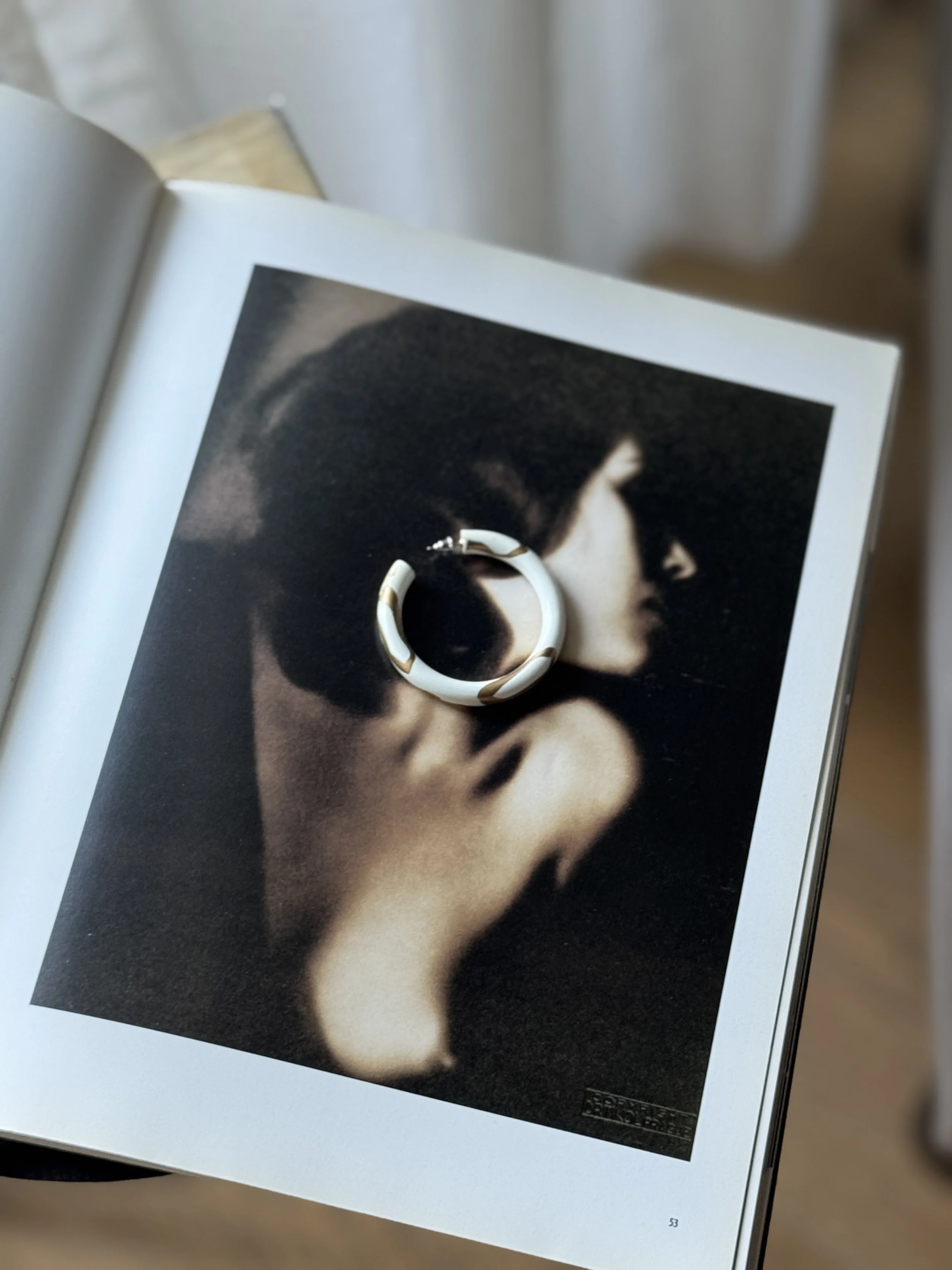An open book with a black-and-white photograph of a woman's profile, wearing large sunglasses, resting on it. A white and gold circular ring is placed on the photo. nude art photography