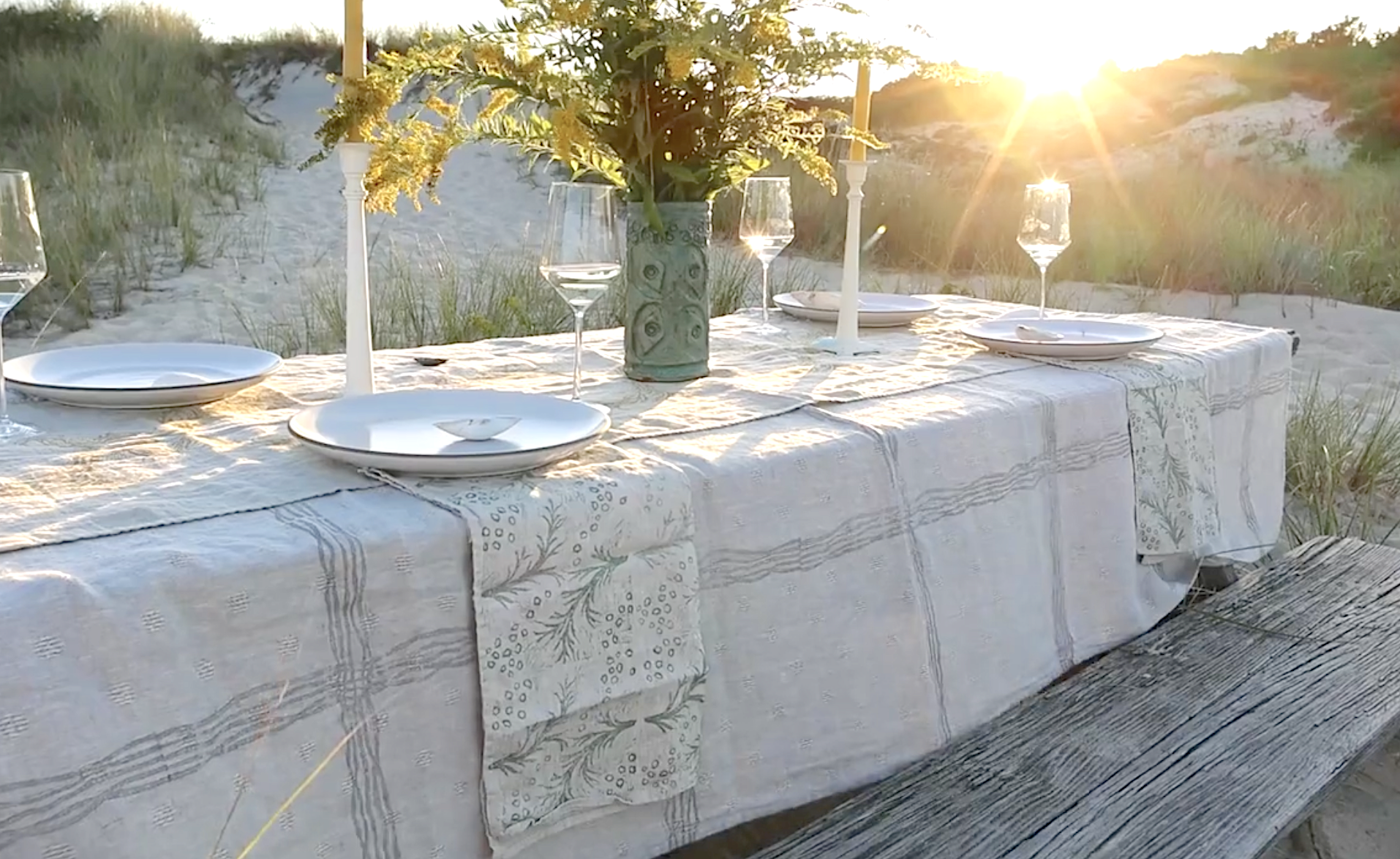 Decorated outdoor dining table on the beach with white plates, wine glasses, a floral centerpiece in a decorative vase, and white candles, set during sunset with beach sand and grass surrounding.