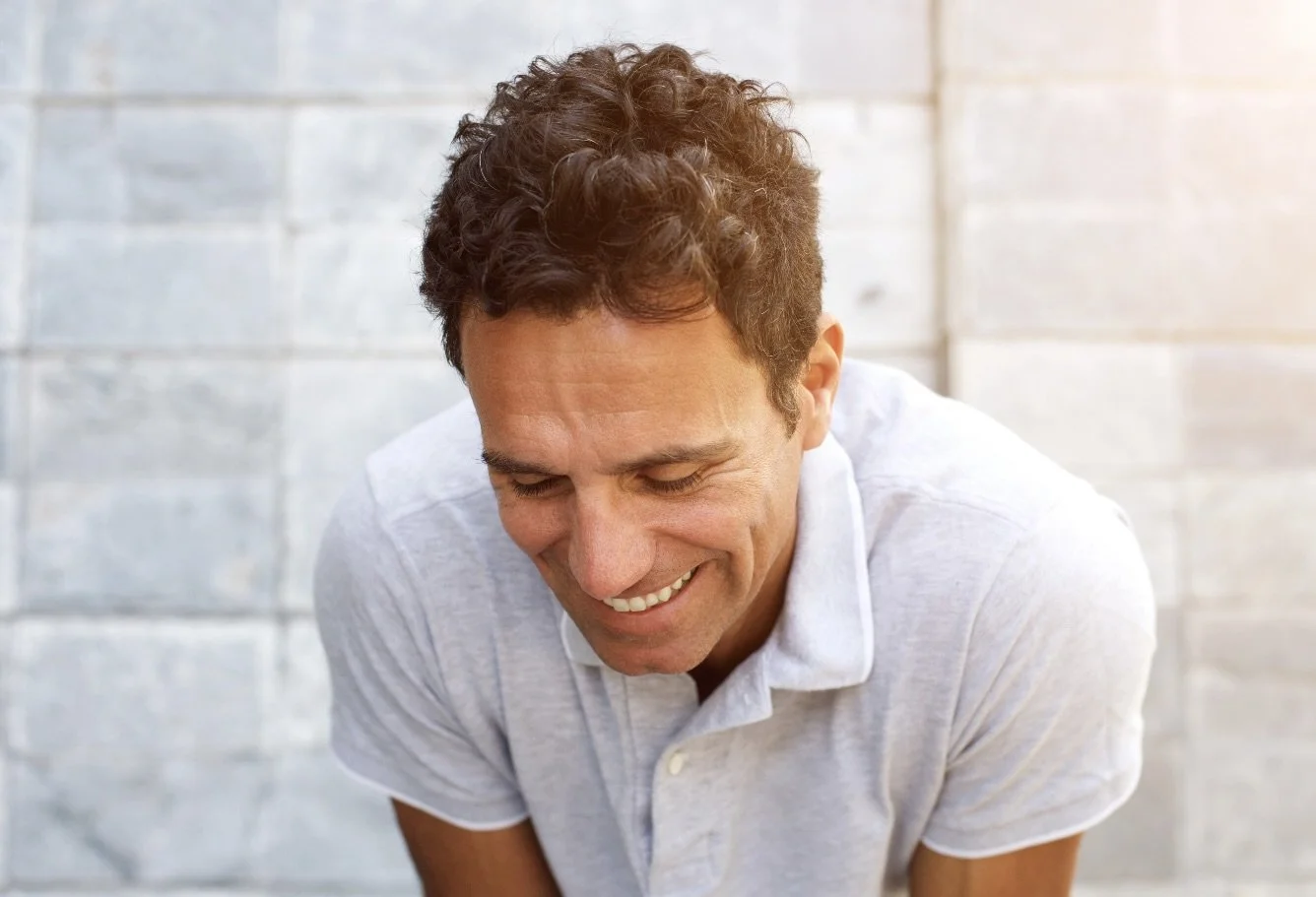 A man with curly brown hair is smiling with his eyes closed, wearing a light gray polo shirt, standing against a light-colored brick wall.