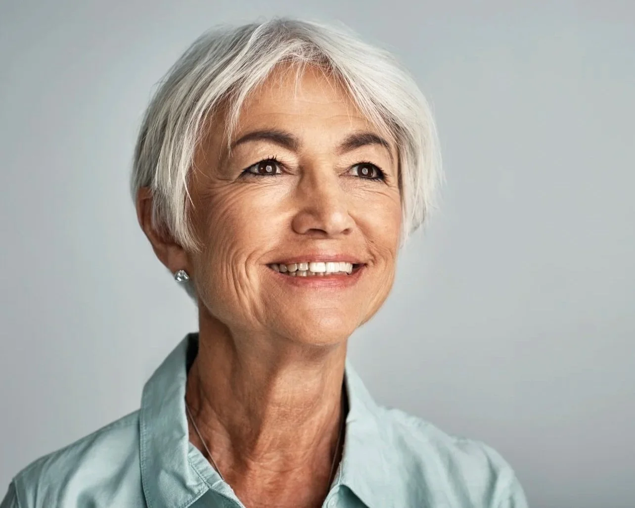 Close-up of a smiling older woman with short gray hair, wearing a light blue collared shirt, against a plain light gray background.