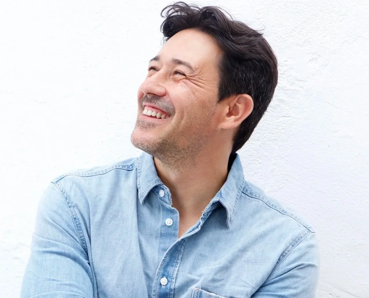 A smiling man with dark hair and light skin, wearing a light blue denim shirt, standing against a white textured wall, looking to the left