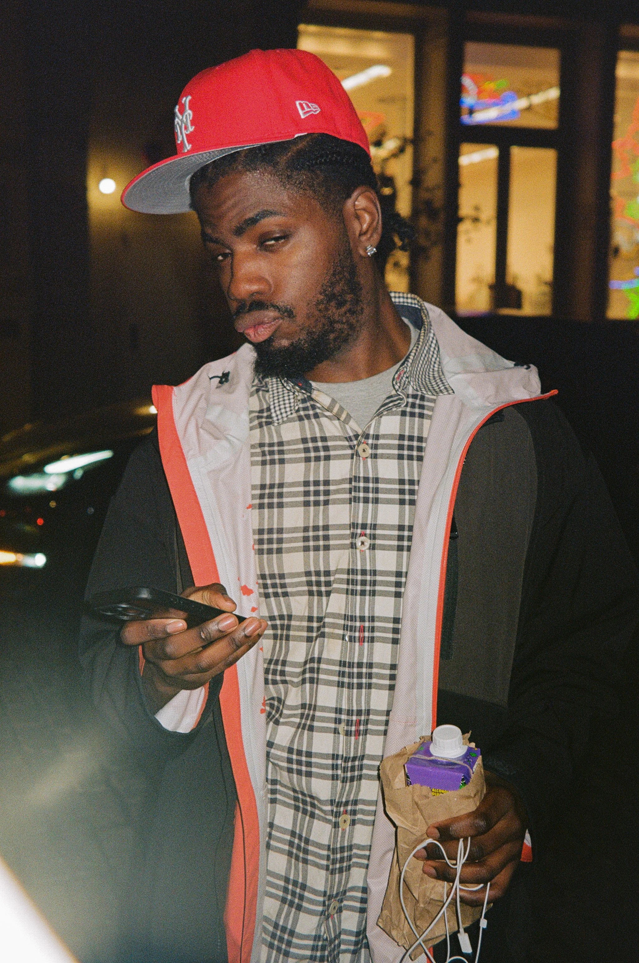 A young man wearing a red baseball cap, plaid shirt, and black jacket, looking at his phone while standing outside at night.