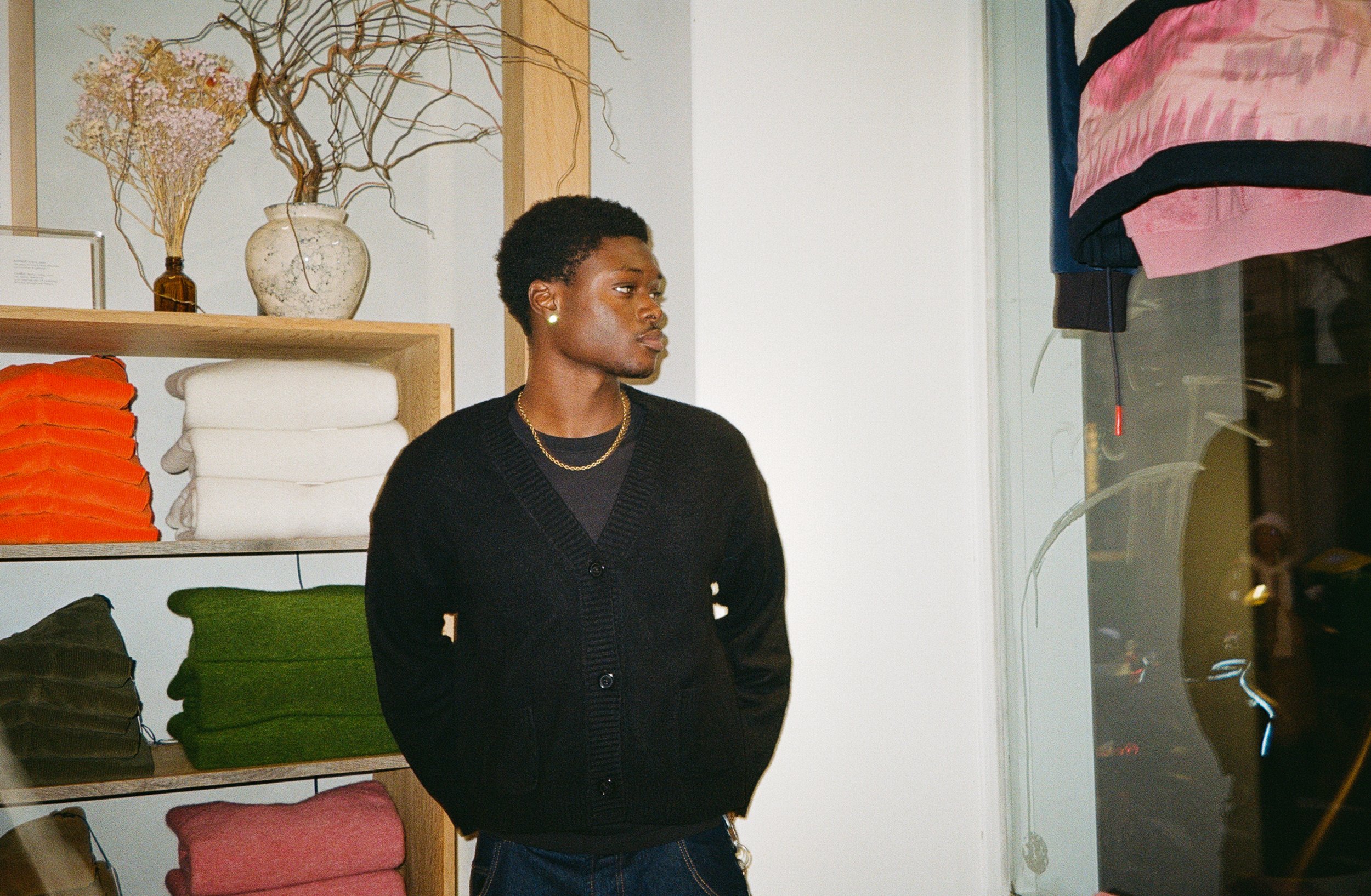 A young man with dark skin and short curly hair stands in a store, wearing a black cardigan, black t-shirt, gold chain, and small pearl earrings. Behind him are shelves with folded fabric or clothing in various colors, and a decorative vase with dried flowers. The store has a modern, minimalist look.
