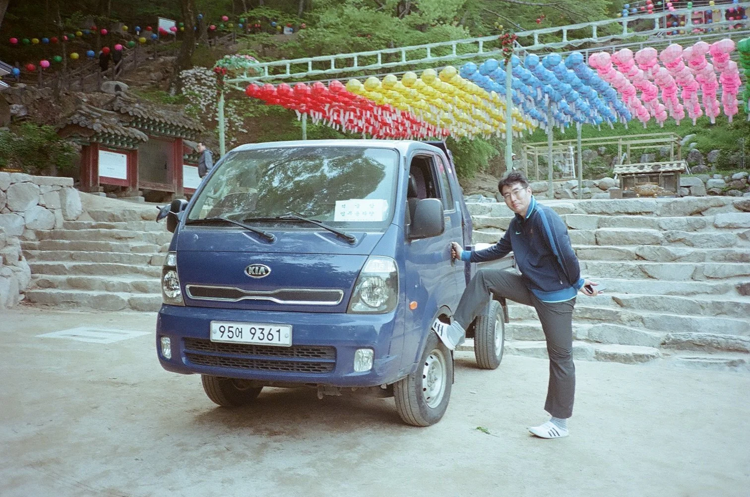 A man in a dark jacket and gray pants standing with one foot on the wheel of a small blue Kia truck, holding the door handle. The background features stone steps, colorful paper lanterns hanging overhead, and a traditional pavilion among trees.