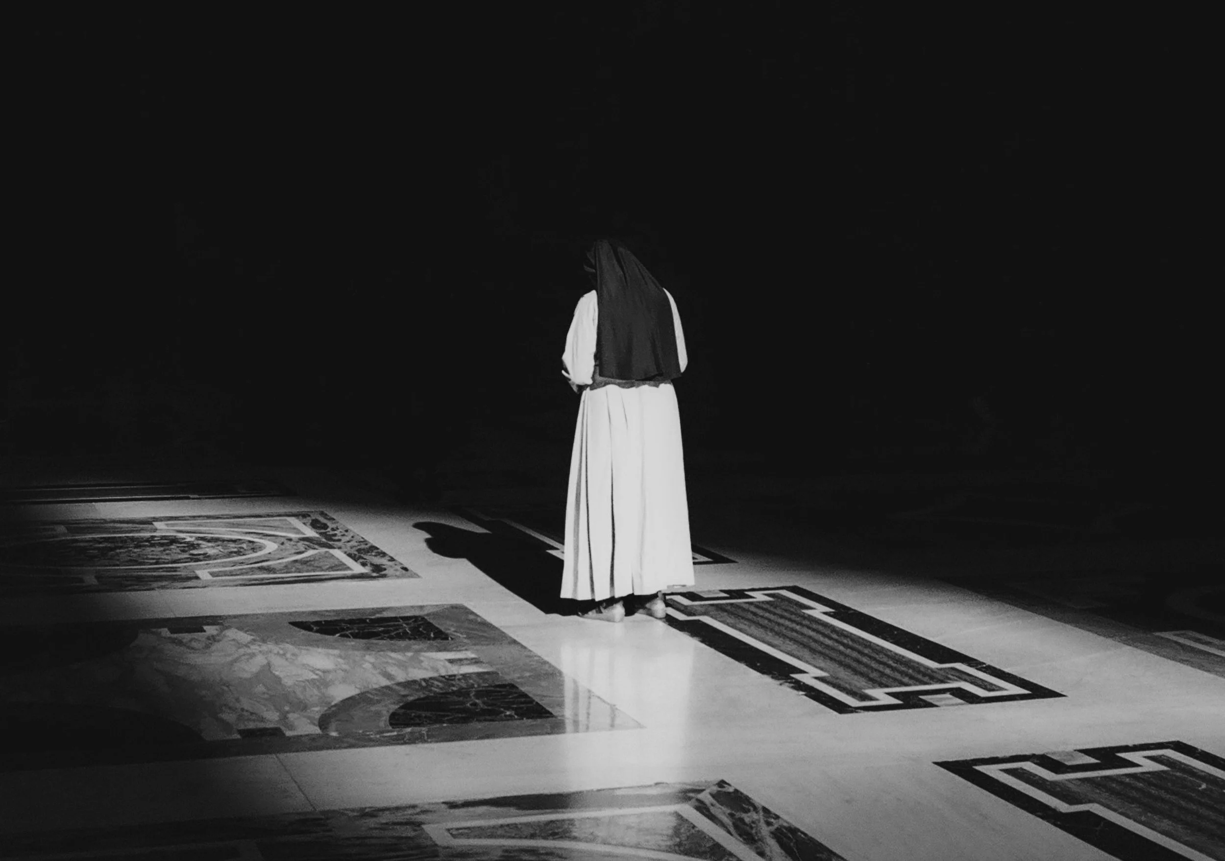 A person standing alone in a dark room on a decorated floor, illuminated by a single light source, with their back turned to the camera.