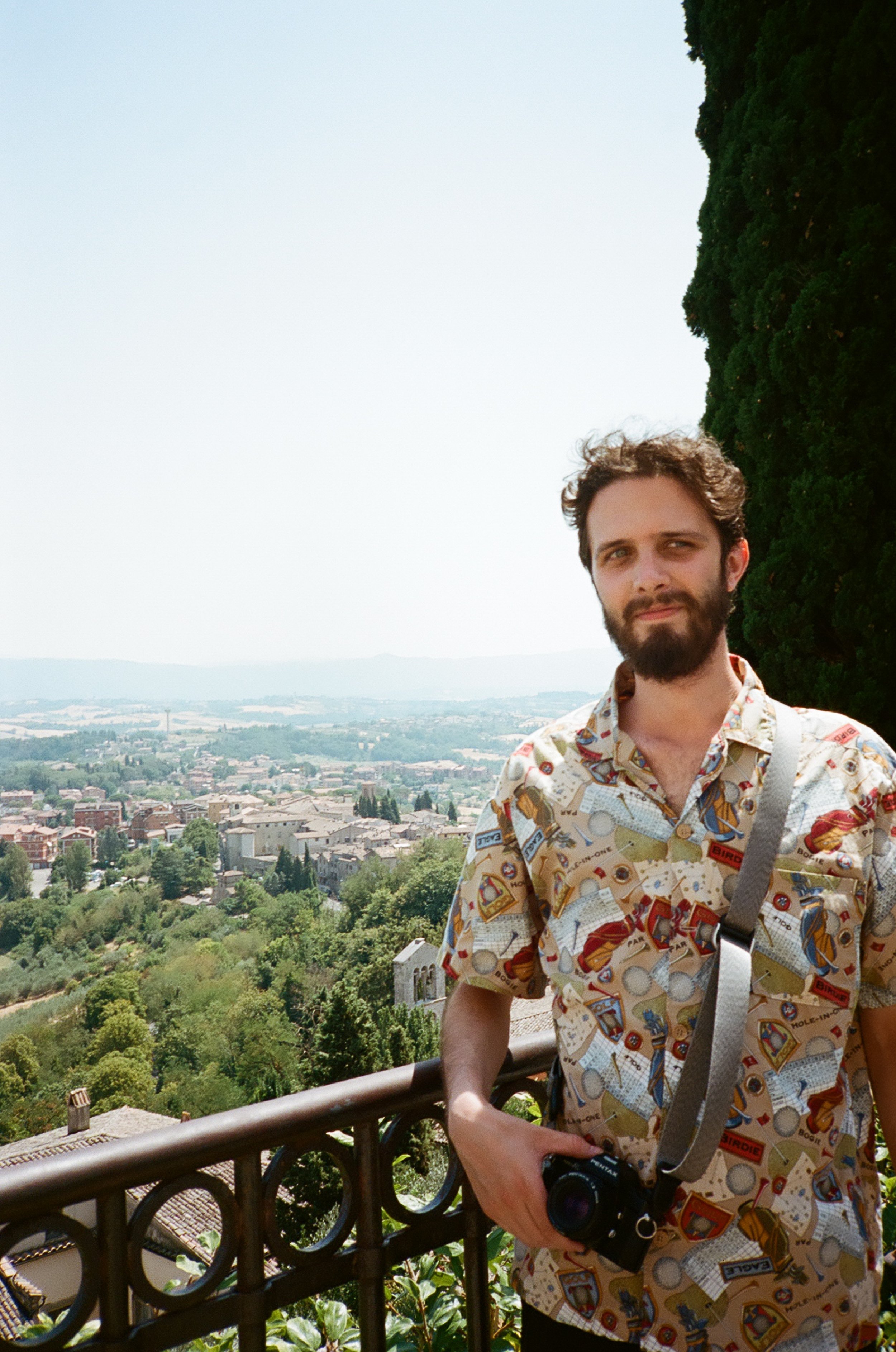 A man with a camera around his neck standing outdoors on a balcony, with a scenic view of a city and green landscape in the background.