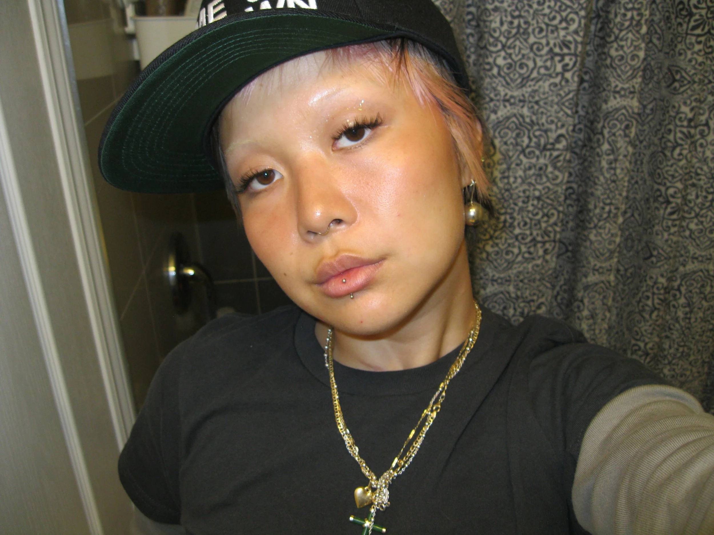 A person taking a selfie in a bathroom, wearing a black cap tilted sideways, multiple necklaces, and a black shirt with long sleeves. They have short, light brown or pinkish hair, and several facial piercings including a septum ring, a labret piercin