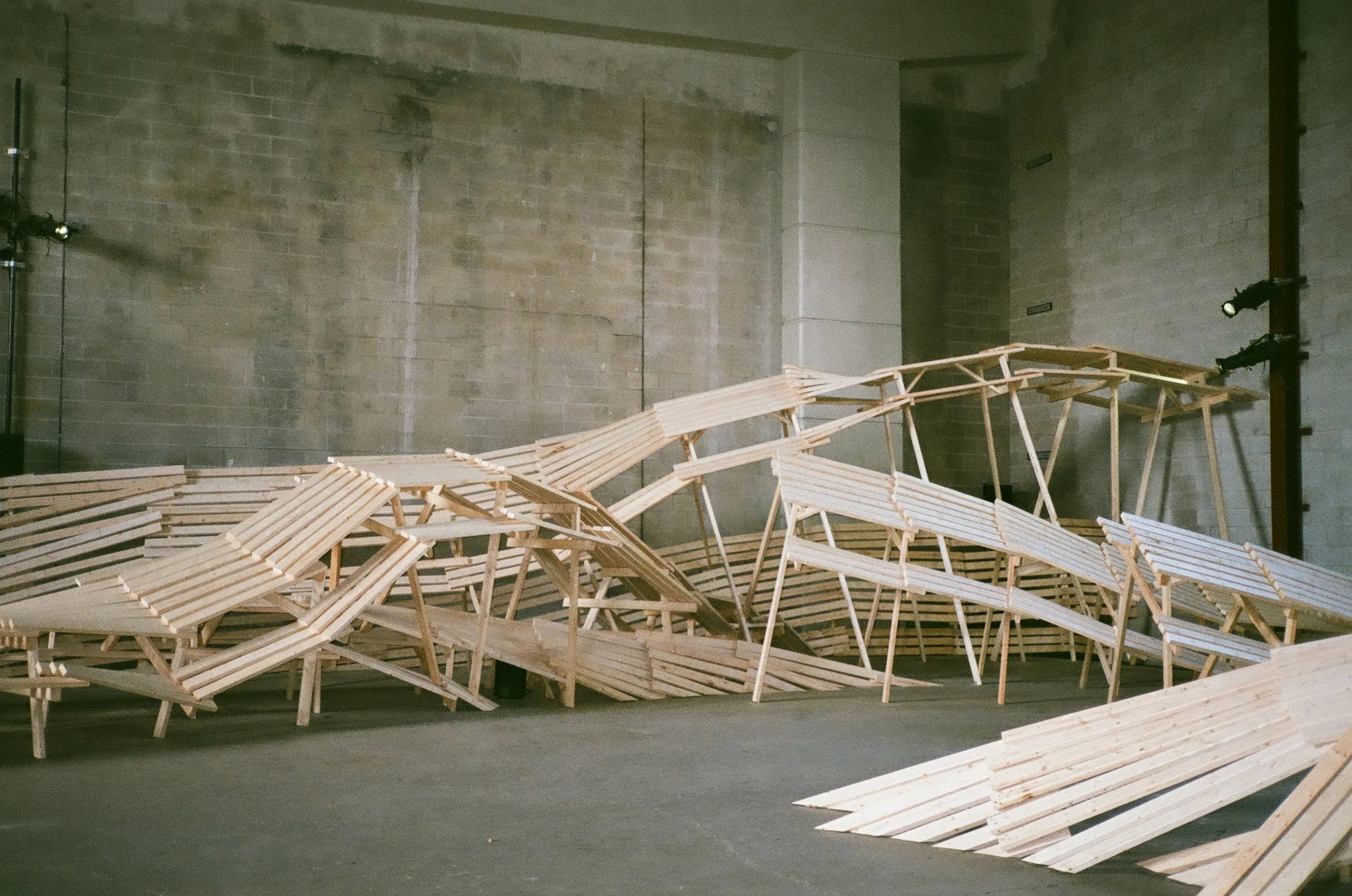 Unfinished wooden installation or sculpture with arranged slats and frames inside an indoor industrial space with brick walls.
