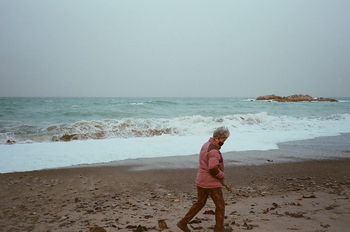 An elderly woman with gray hair, wearing a face mask, pink jacket, and brown pants, walking on a rocky beach near the ocean during cloudy weather.