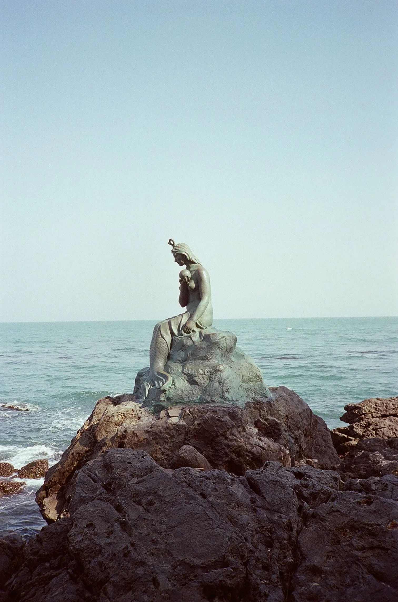 Statue of a mermaid sitting on rocks by the ocean with a clear sky in the background.