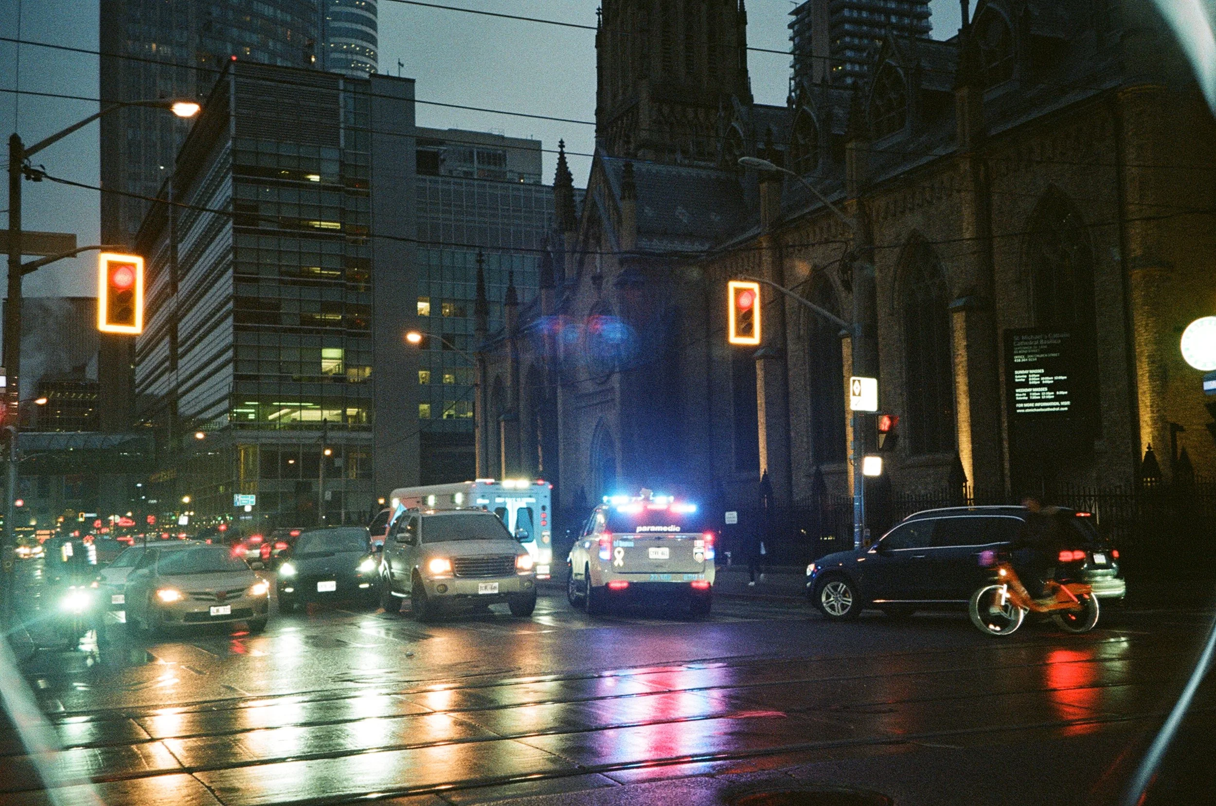 A city street at dusk with a church, modern buildings, cars, a police car with flashing lights, an ambulance, and a cyclist.