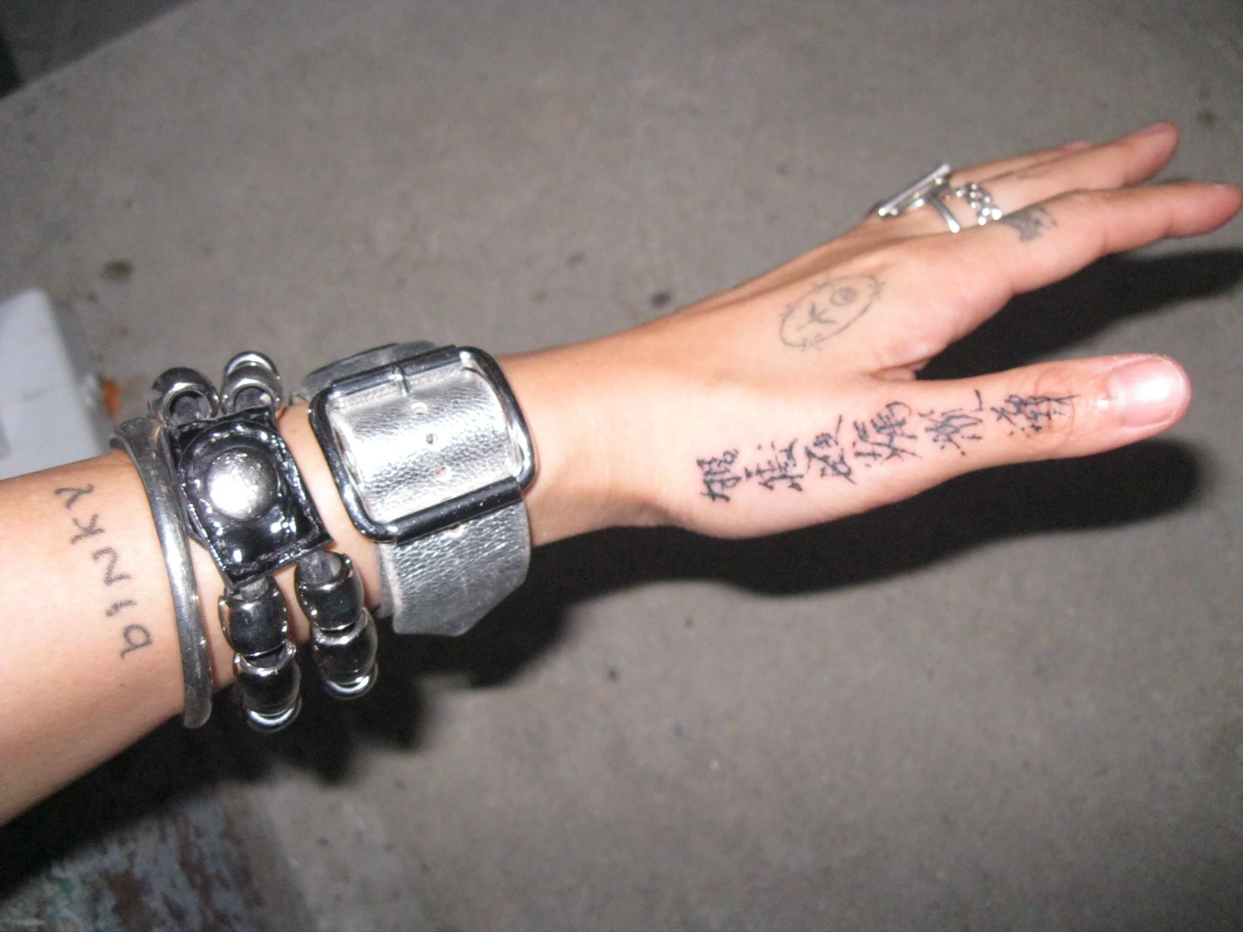 A person's hand and wrist with multiple rings and bracelets, tattoos, and writing on the fingers, against a gray background.