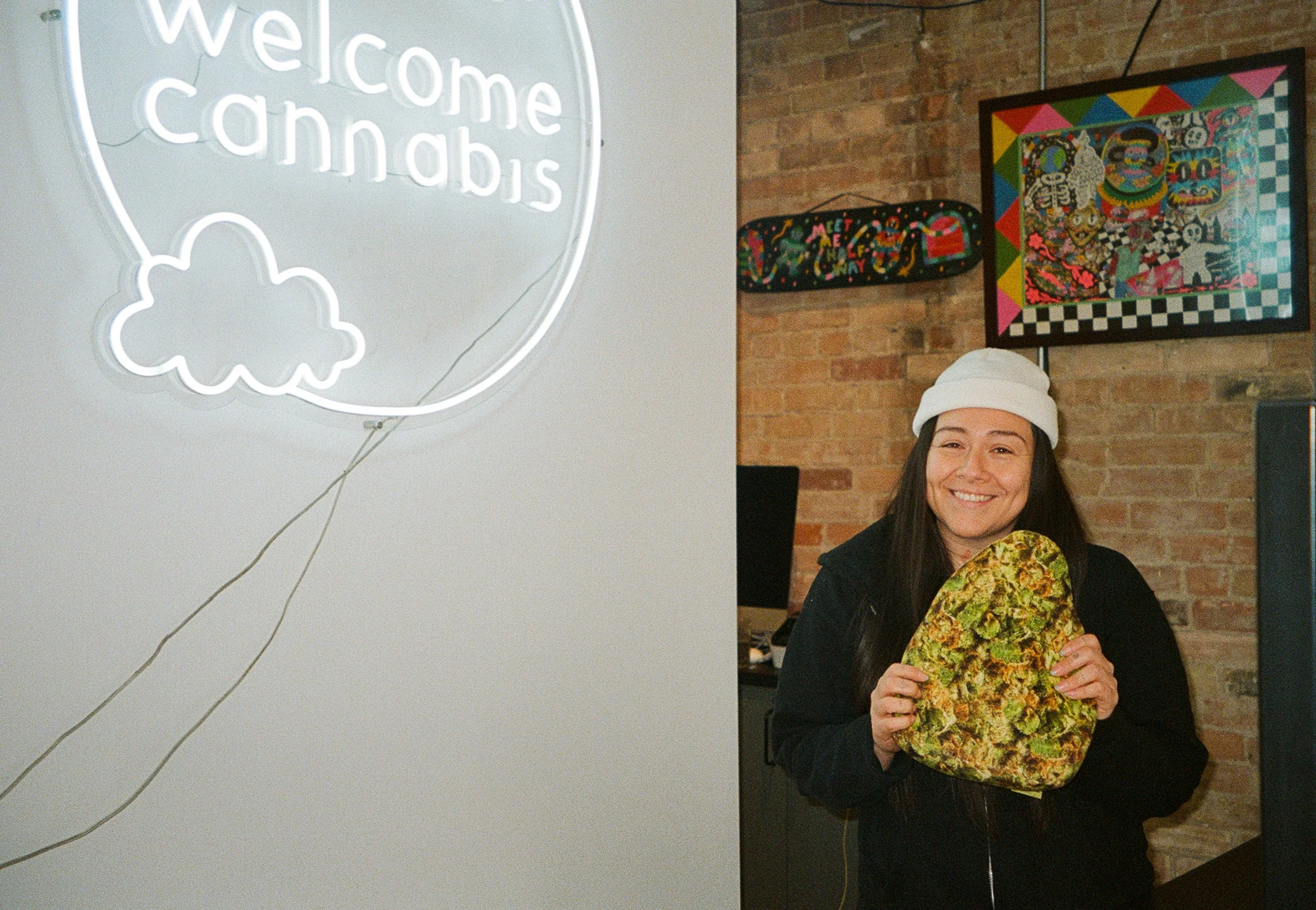 Person with long dark hair smiling, wearing a white beanie, holding a large, colorful marijuana bud, standing inside a brick-walled room with vibrant artwork and a neon sign that says 'Welcome Cannabis'.