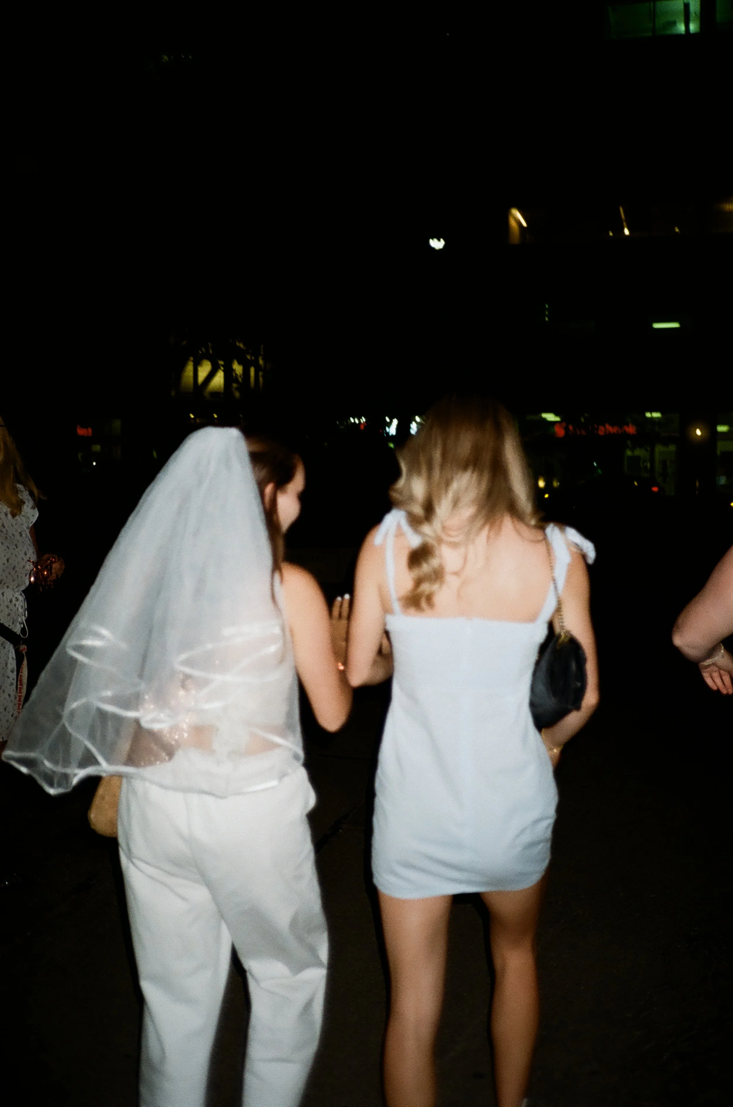 Two women walking together at night; one wears a white dress with bows on the shoulders and a veil, the other wears a white sleeveless dress with a black purse.