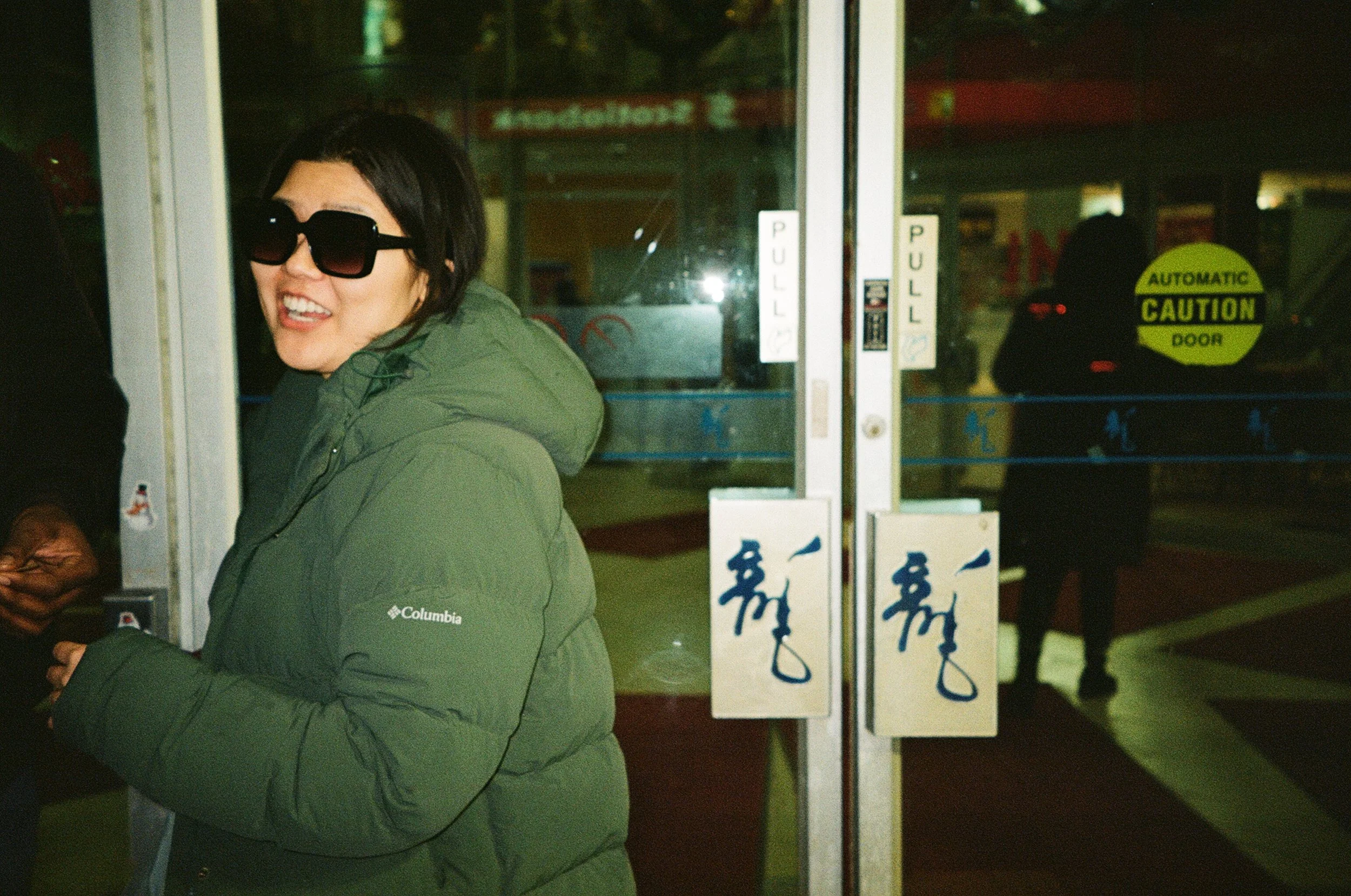 A woman wearing a green Columbia winter jacket and large black sunglasses standing at the entrance of a building, holding an object in her hands, with glass doors behind her.