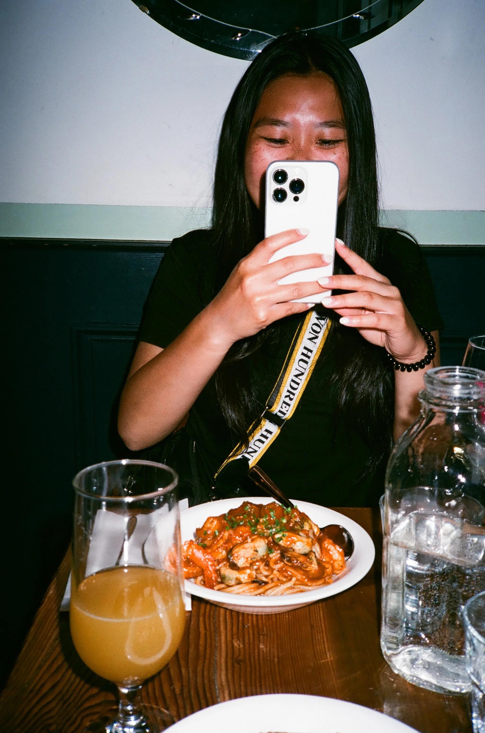 A woman with long black hair taking a selfie with a white smartphone at a dining table. In front of her is a plate of seafood pasta and a glass of orange juice, with a pitcher of water nearby.