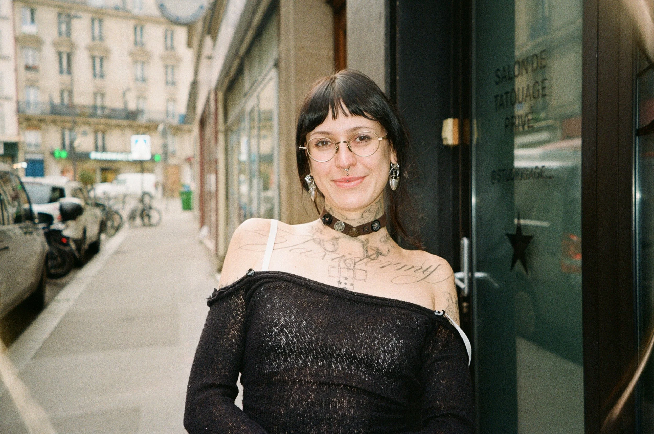A woman with black hair and glasses stands on a city sidewalk outside a tattoo studio, smiling at the camera.