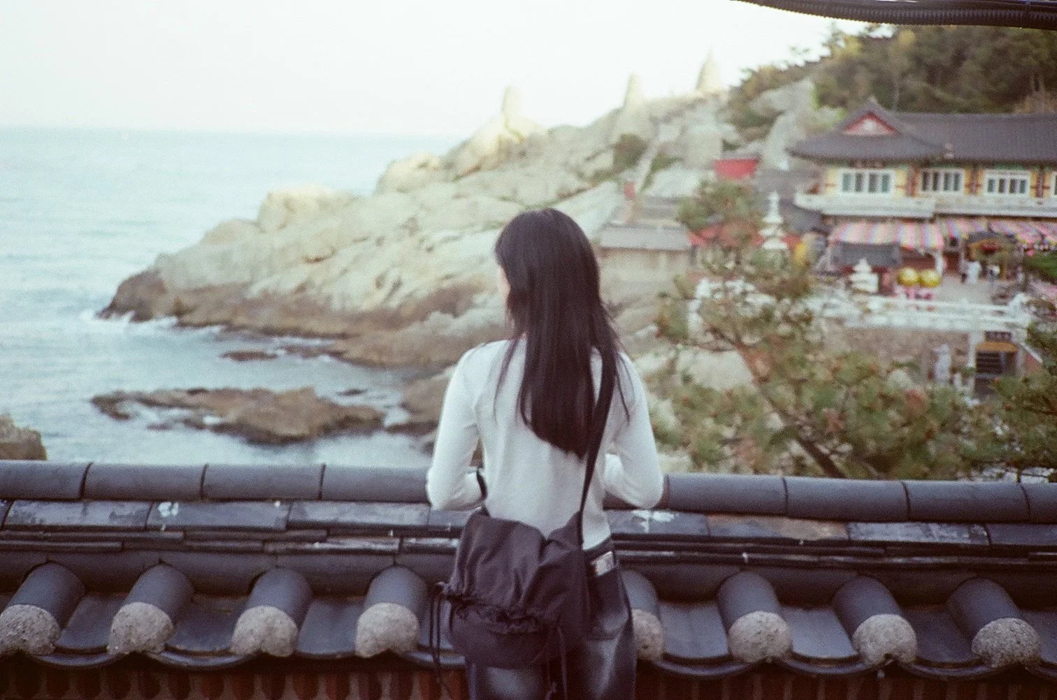 A woman with long black hair, dressed in a white top and black pants, stands on a balcony with traditional Asian roof tiles, looking out at a coastal landscape with rocky cliffs, the ocean, and a traditional building on a hill in the distance.