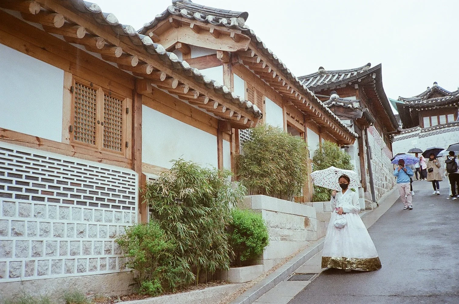 Woman wearing a white dress and black face mask holding a white umbrella with a floral pattern, standing on a city street with traditional Asian architecture, other people with umbrellas walking in the background.