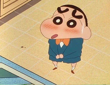 Cartoon boy with big eyes and blushing cheeks, standing with hands clasped, looking up with a nervous expression.